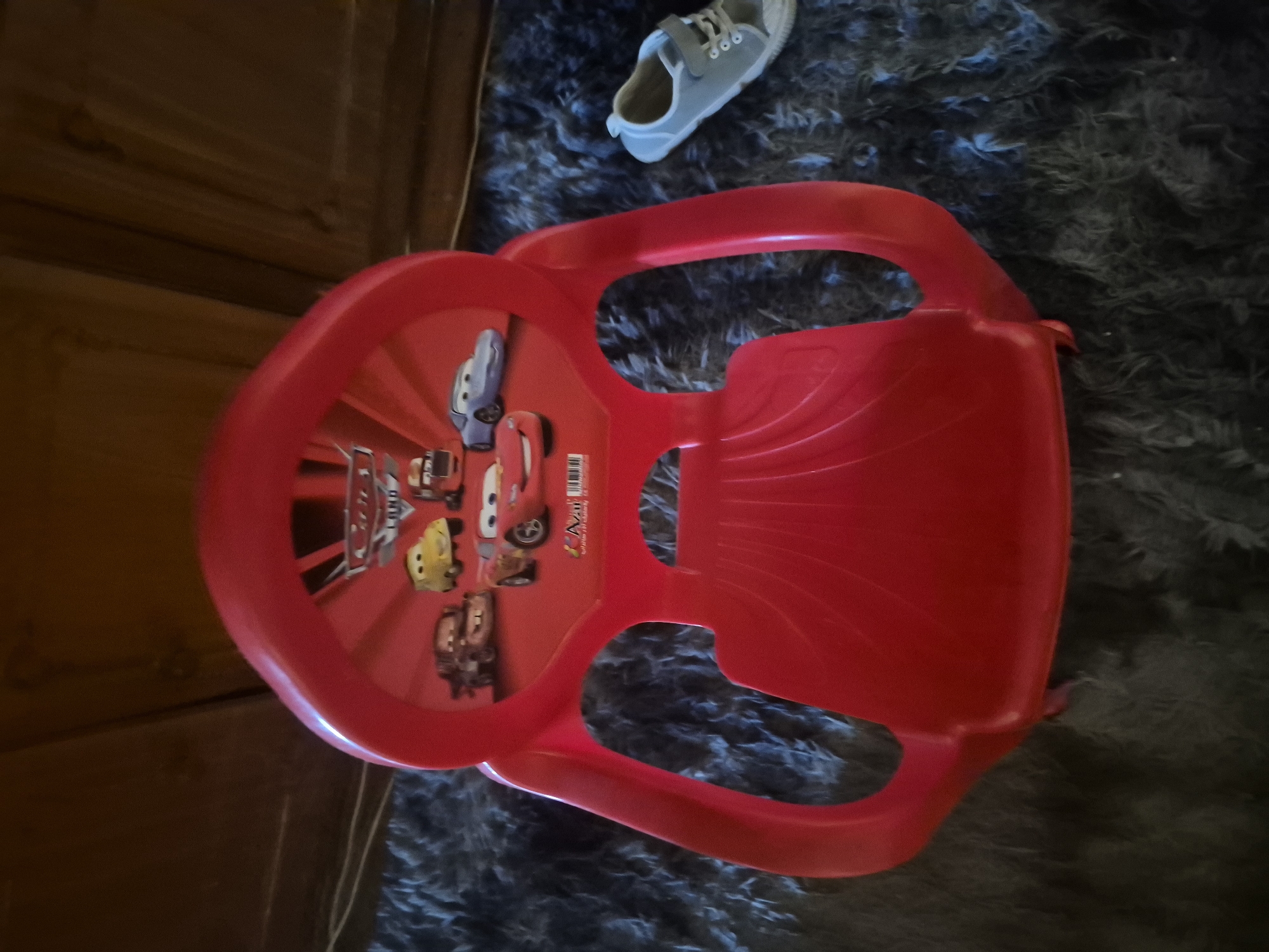 A red plastic children's toy sled, decorated with images from the Disney/Pixar movie "Cars," is positioned on a dark, fuzzy rug. To the right of the sled, a single grey child's sneaker with white laces and a velcro strap is visible on the same rug. The background consists of a wooden surface, likely furniture, and a dark, textured wall. The scene appears to be indoors, within a residential setting. There is no indication of the city of Yerevan, Armenia, or any specific location.