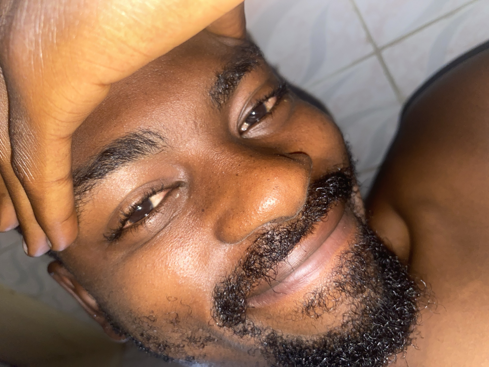 This media file presents a close-up portrait of a Black man, likely from Jos, Nigeria, focusing on his face and upper body. He appears to be an adult in his 20s or 30s, characterized by his dark brown skin, full dark beard, and mustache. The man is positioned with his head tilted towards the upper left of the frame, looking directly into the camera with a relaxed and gentle smile. His dark brown eyes are expressive, reflecting a soft light, and conveying a friendly and approachable demeanor.
His right hand is casually placed over his forehead and temple, partially obscuring the upper left portion of his face. Notable details include the clear and healthy appearance of his skin, which exhibits a slight natural sheen. His beard is thick and features dense, dark, curly hairs. The background is softly out of focus but reveals light-colored tiles with visible grout lines, suggesting an indoor setting, possibly a bathroom or a room with tiled walls. The lighting is soft and even, highlighting his facial features without harsh shadows. There is no visible text in the image. The overall impression is one of quiet contentment and warmth.