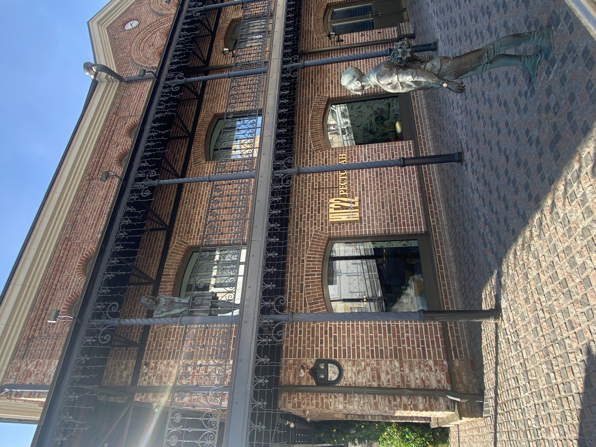 The image shows a brick building with a black metal balcony in Kazan, Russia. A bronze sculpture of a man holding flowers stands on the right. The building has arched windows, and a sign reads "PECTOPAH." A black metal address plaque is visible on the wall. The sidewalk is paved with grey cobblestones. The sky is blue.