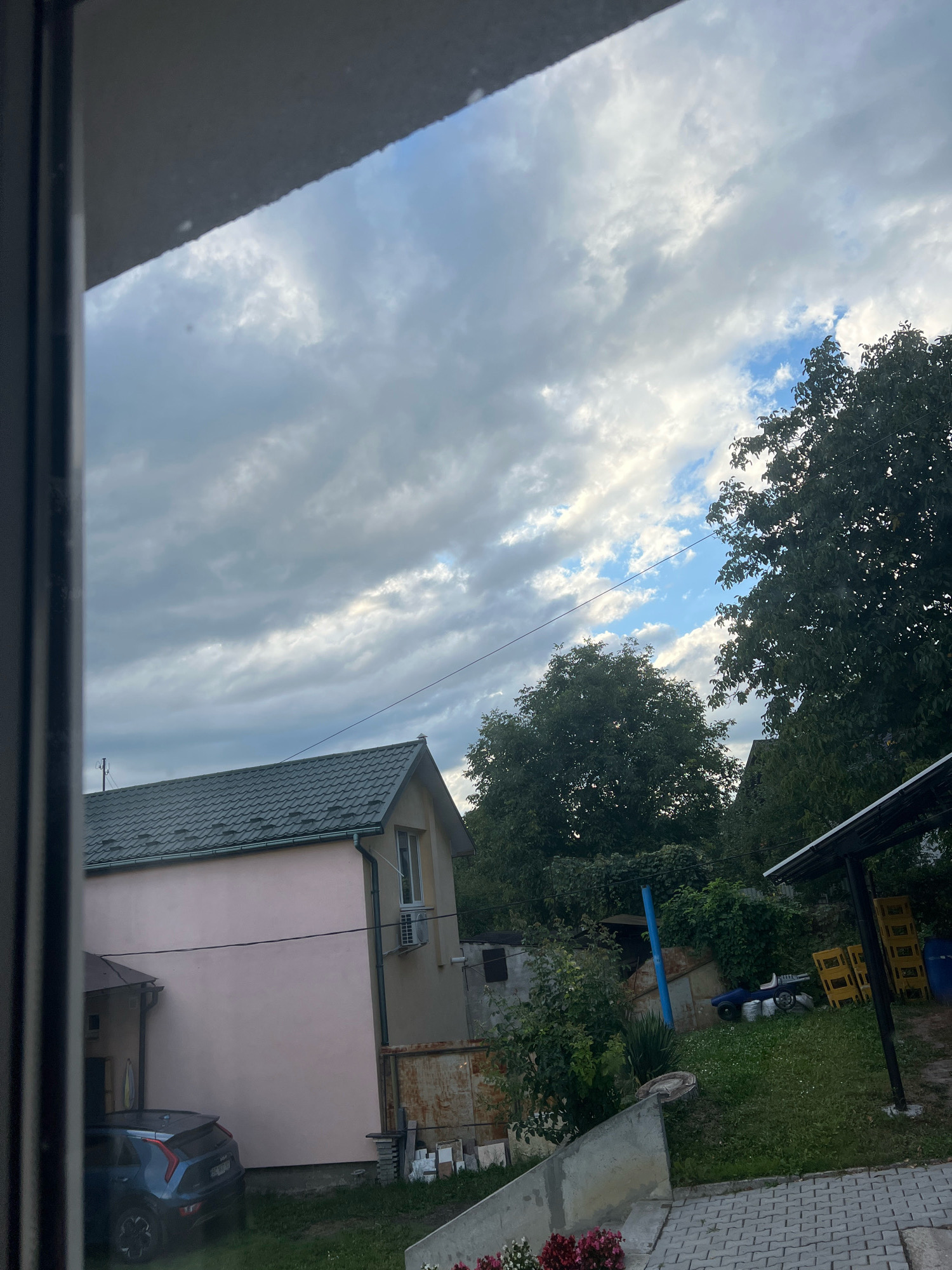 This is a photograph taken from a window, looking out onto a residential backyard in Drohobych, Ukraine. The sky is cloudy with patches of blue visible. In the foreground, on the left, is the edge of the window frame.
The middle ground of the image features a pale pink, two-story house with a dark green, tiled roof. There is a window and an air conditioning unit visible on the side of the house. To the left of this house is a darker structure, possibly an attached garage or extension. A dark blue hatchback car is parked in front of this structure.
The background shows dense green trees. To the right, there is a covered area with a black roof, supported by posts. Yellow plastic crates and a blue barrel are stacked underneath this shelter. The ground is covered in grass, and a concrete walkway made of interlocking pavers is visible in the bottom right corner of the image. Some potted flowers are also visible in the lower left corner. The overall impression is that of a slightly overcast day in a suburban or rural setting.