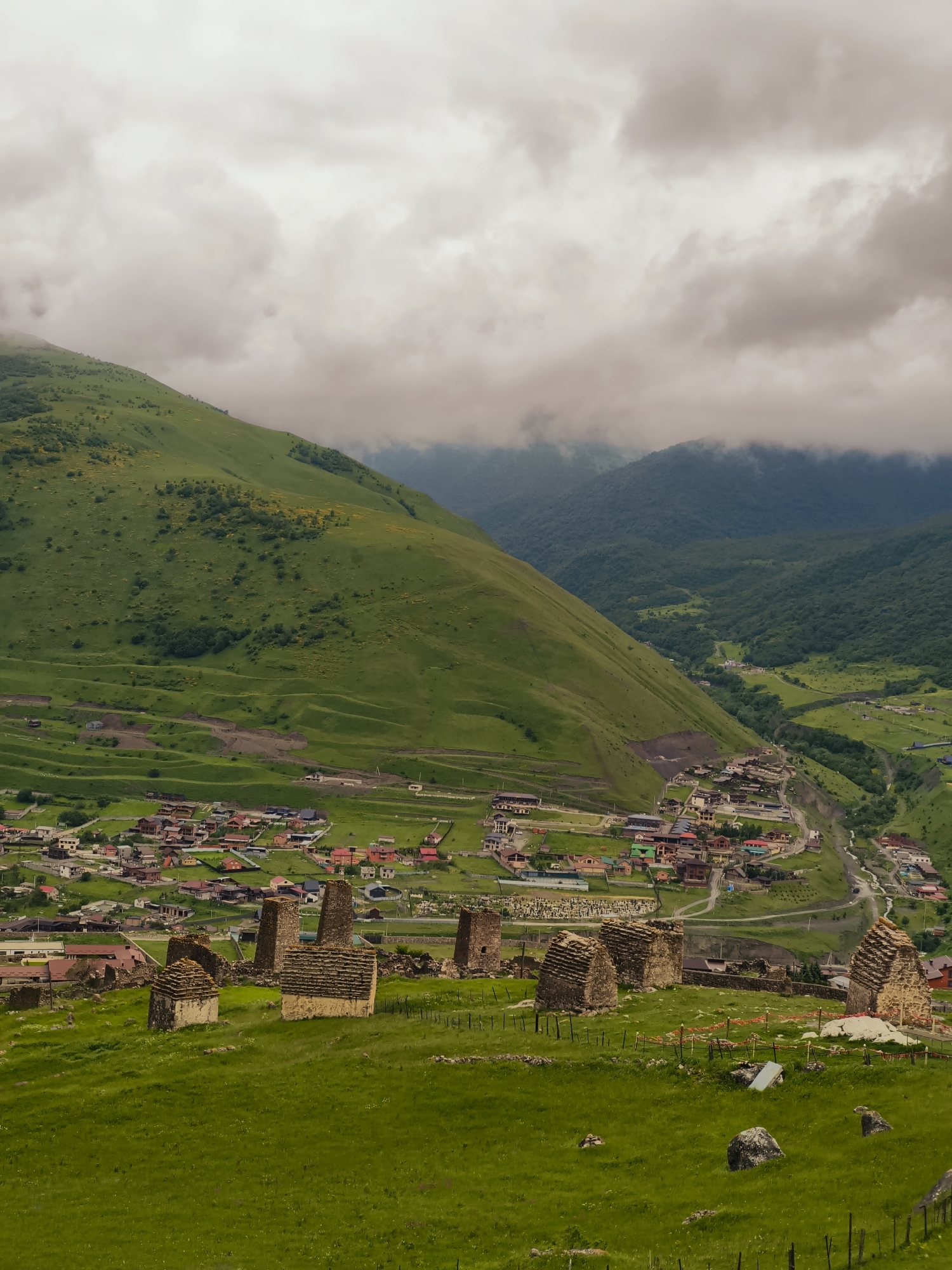 The image displays a panoramic view of the city of Verkhny Fiagdon, Russia, nestled within a lush green valley. In the foreground, ancient stone crypts, some appearing as stone towers and others as smaller, hut-like structures with layered roofs, are scattered across a grassy hill. A low fence with wooden posts demarcates the area around these historical structures.
Beyond the crypts, a picturesque village unfolds with numerous houses featuring red, brown, and grey roofs, clustered together and connected by winding roads. The village extends down the gentle slope of a large, verdant mountain that dominates the left side of the frame. This mountain is characterized by terraced fields and patches of yellow wildflowers.
Further into the distance, the valley narrows, revealing a winding river and more dense, tree-covered mountains shrouded in a soft, overcast sky. The clouds are thick and grey, casting a diffused light over the entire landscape. There are no people visible in the image, and the scene is static and serene, capturing a historical and natural environment. The overall impression is one of timelessness and integration with the surrounding natural beauty.