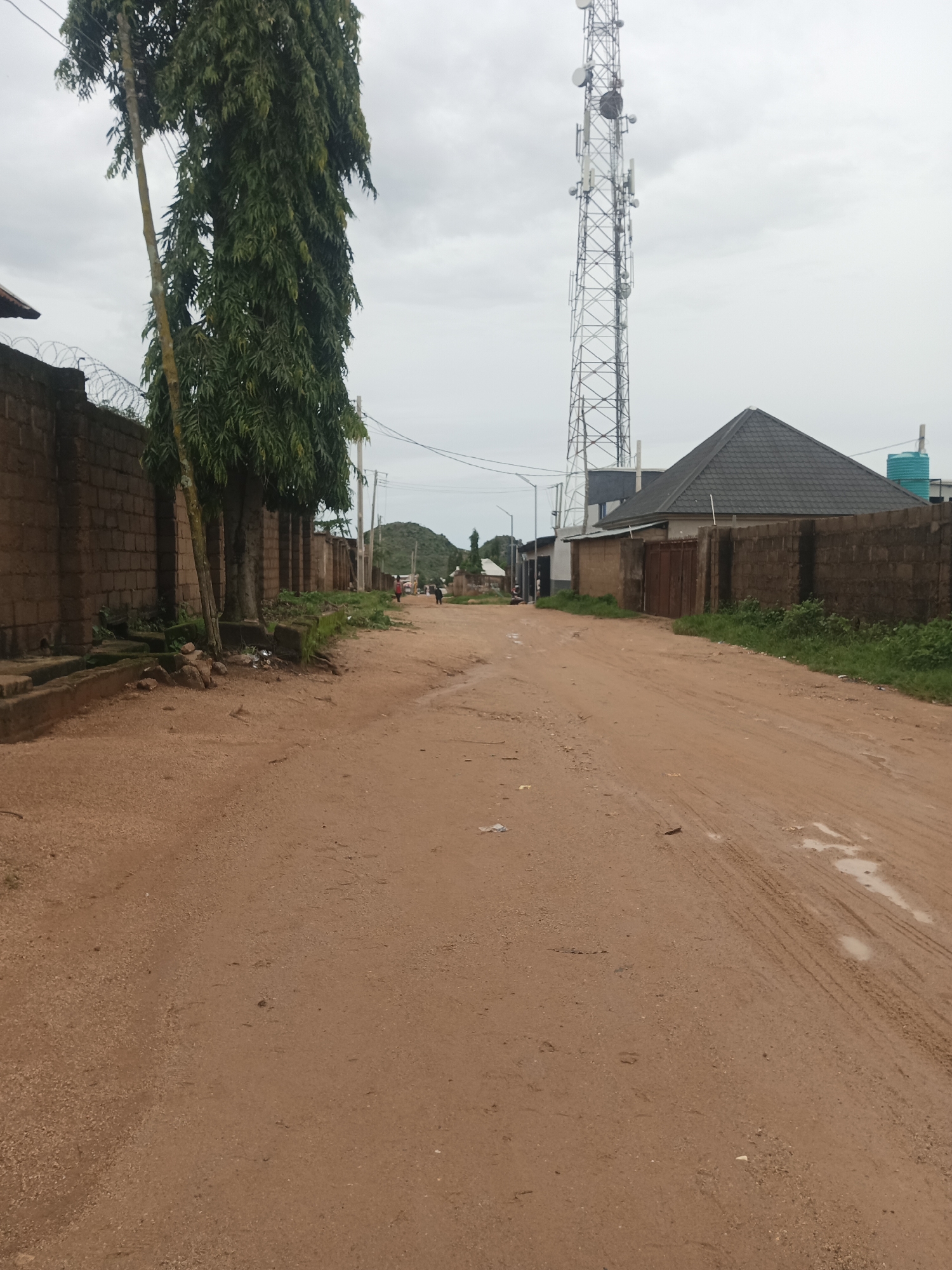 The image depicts a dirt road in Jos, Nigeria. On the left side of the road, a brick wall topped with barbed wire runs along the property line. Several tall trees with dense green foliage are situated in front of the wall. A cell tower is visible in the background, to the right of the trees. Further down the road, on the right side, a building with a dark, sloped roof and a brick wall encloses the property. Several people are walking and standing in the distance on the road, and a motorbike is parked on the left side of the road. The road is unpaved and has some puddles, indicating recent rainfall. The sky is overcast.