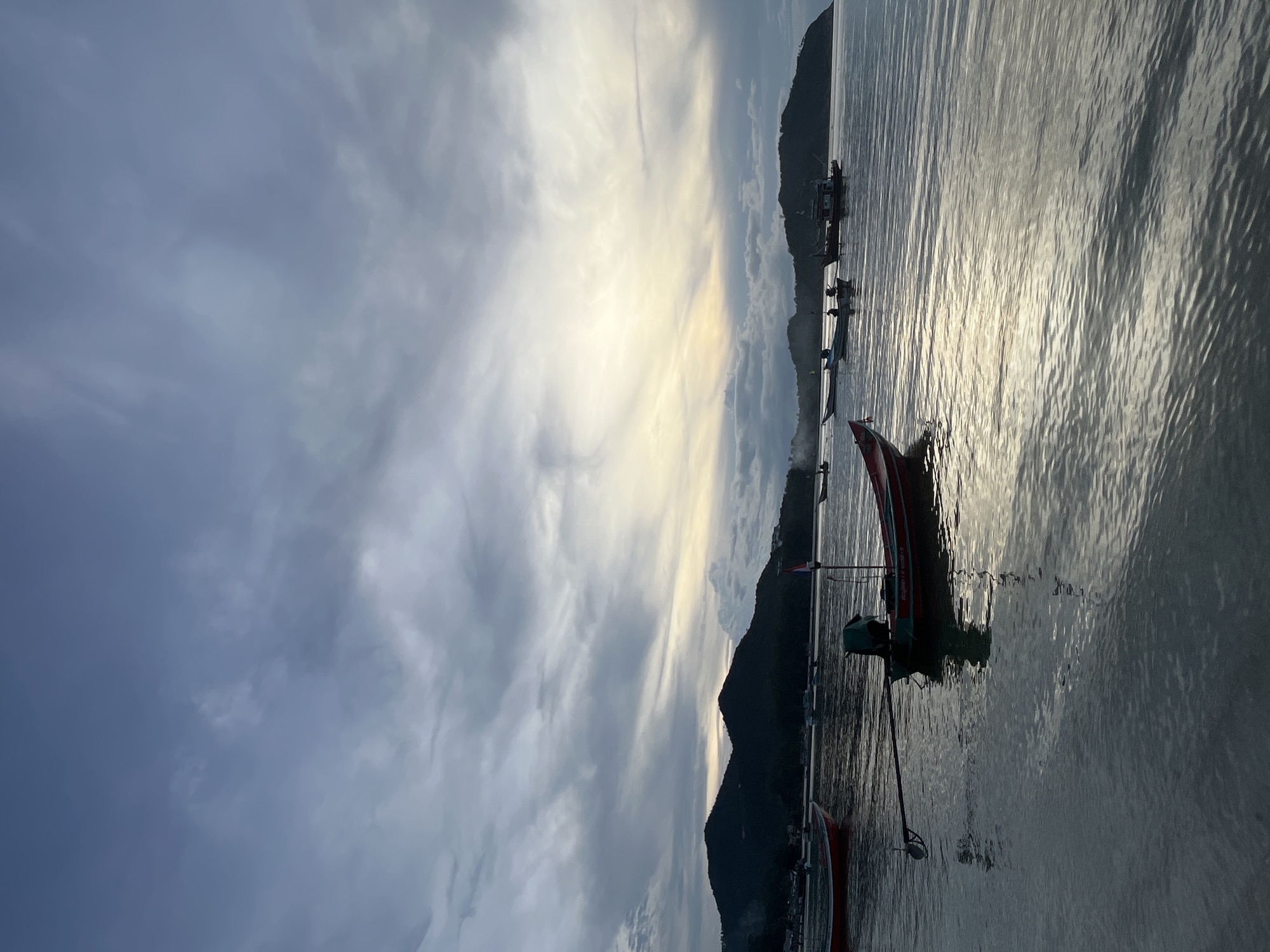 The image captures a coastal scene in ตำบลเกาะพะงัน, Thailand, under a largely overcast sky. The upper portion of the frame is dominated by dense, grey, and blue clouds, with a discernible band of soft, yellowish light appearing near the horizon on the right, indicating a low-angle sun obscured by clouds. A chain of dark, silhouetted mountains or large hills spans the mid-ground, with faint architectural structures visible on their slopes. The water in the foreground and mid-ground is relatively calm, displaying reflections of the sky's light and cloud patterns.
Several boats are distributed across the water. A prominent red and green longtail boat, characterized by its extended propeller shaft, a green tarp or covering near the engine, and a small flag on a pole at the stern, is positioned in the lower-middle of the frame. The bow of another red boat is visible on the far left. To the left of the central longtail boat, a lighter-colored boat contains what appears to be one or two indistinct human figures. Further into the mid-ground, additional boats, including a larger dark-hulled vessel, are visible, some moored closer to the distant landmasses. No direct interactions between individuals or boats are discernible.