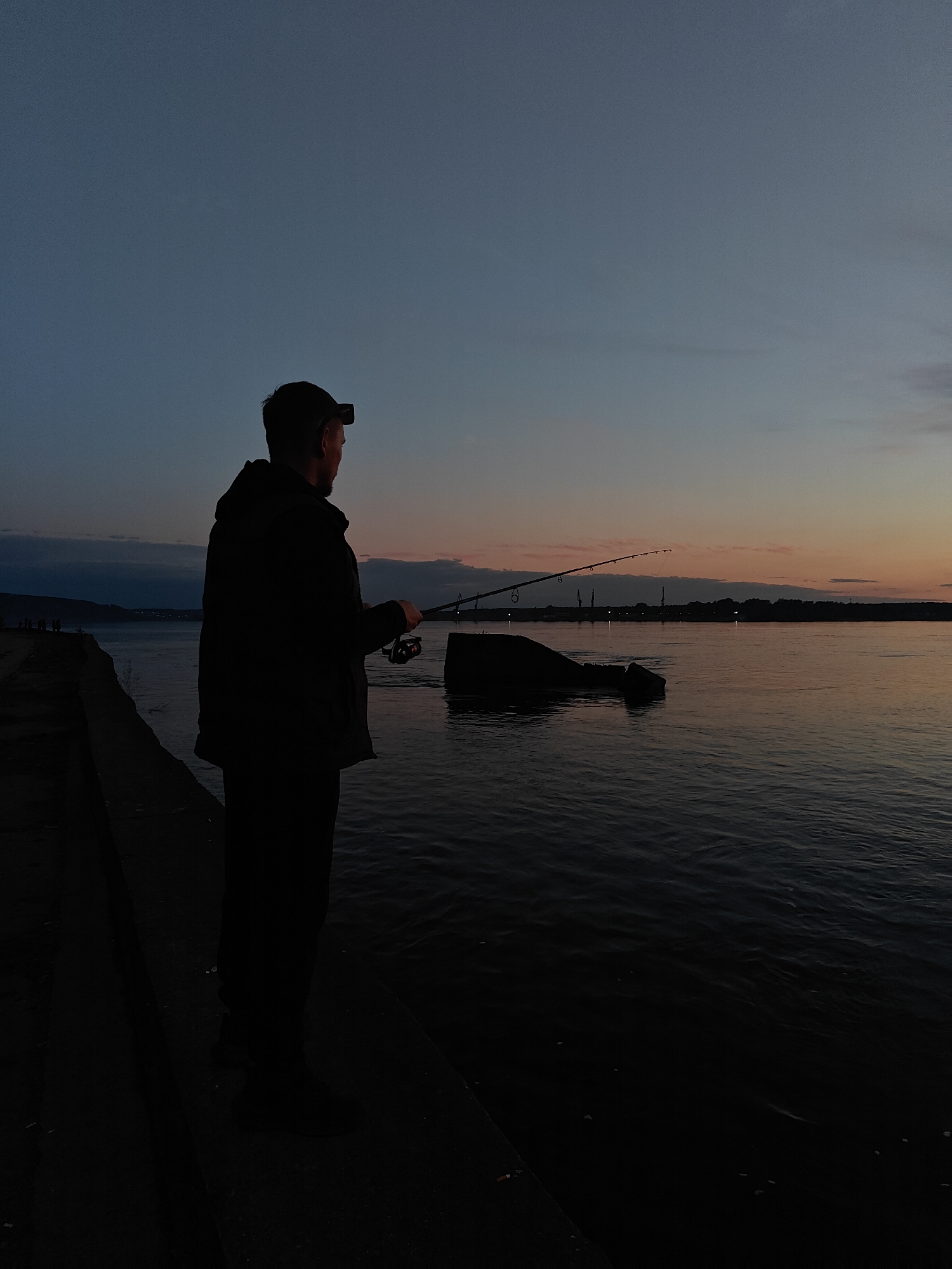 A person is fishing from a concrete embankment on the Kama River in Chaikovsky, Russia. The person is silhouetted against the dusk sky and the river, holding a fishing rod. A headlamp is visible on their head. The river reflects the colors of the sunset. In the distance, the skyline of Chaikovsky can be discerned. A stone breakwater is visible in the river.