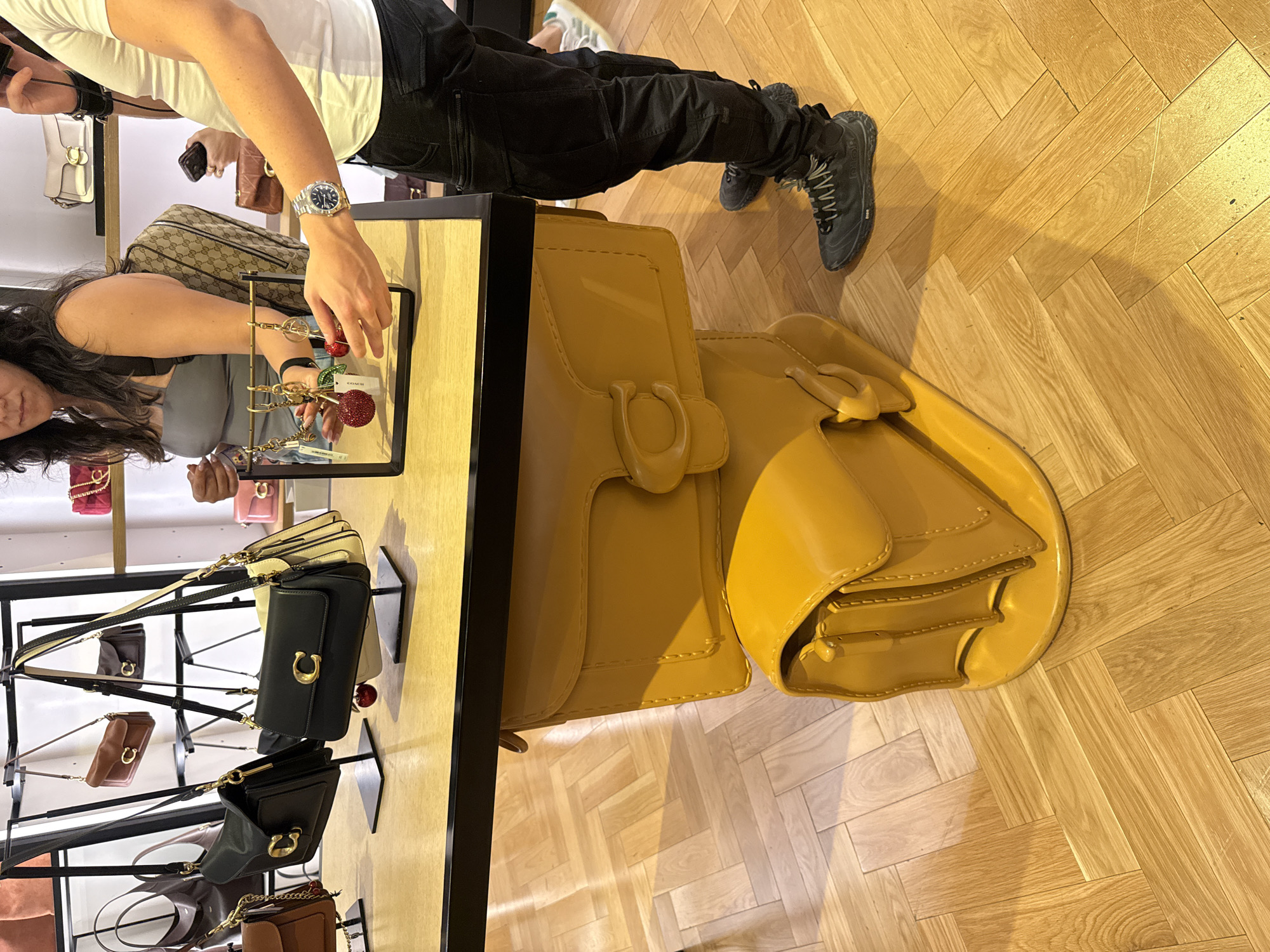 The image depicts the interior of a retail space in Sunrise, Florida, United States of America, likely a fashion accessories store. A person wearing a white t-shirt, dark trousers, and sneakers is standing near a display table. On the table are multiple handbags, including a large yellow bag prominently displayed, along with other handbags on a tiered display. A person with dark hair wearing a grey top is also visible near the table, apparently looking at a display stand with shiny keychains. The floor is wooden and arranged in a parquet pattern.