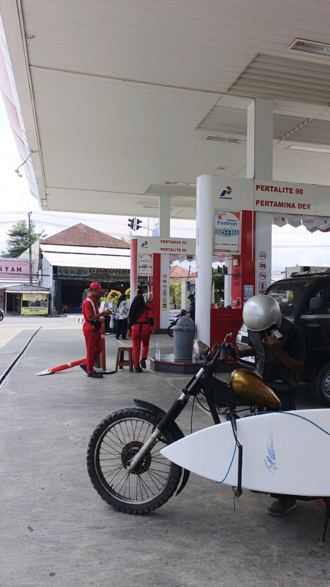 The image depicts a scene at a Pertamina gas station in Desa Wonoharjo, Indonesia. In the foreground, a motorcycle is parked with a white surfboard strapped to its side. A person wearing a silver helmet, dark vest, and blue jeans is interacting with their phone, positioned near the motorcycle.
Behind the motorcycle, a man in a red uniform, likely a gas station attendant, stands next to a red cone. Another person in a similar red uniform, wearing a black headscarf, walks away from the viewer. Several other individuals, some appearing to be customers, are gathered near the gas pumps. The gas station has a white roof with red and white signage indicating fuel types like "PERTAMAX 92," "PERTALITE 90," and "PERTAMINA DEX." A dark-colored SUV is parked at one of the pumps. The environment appears to be an outdoor, paved area typical of a gas station.