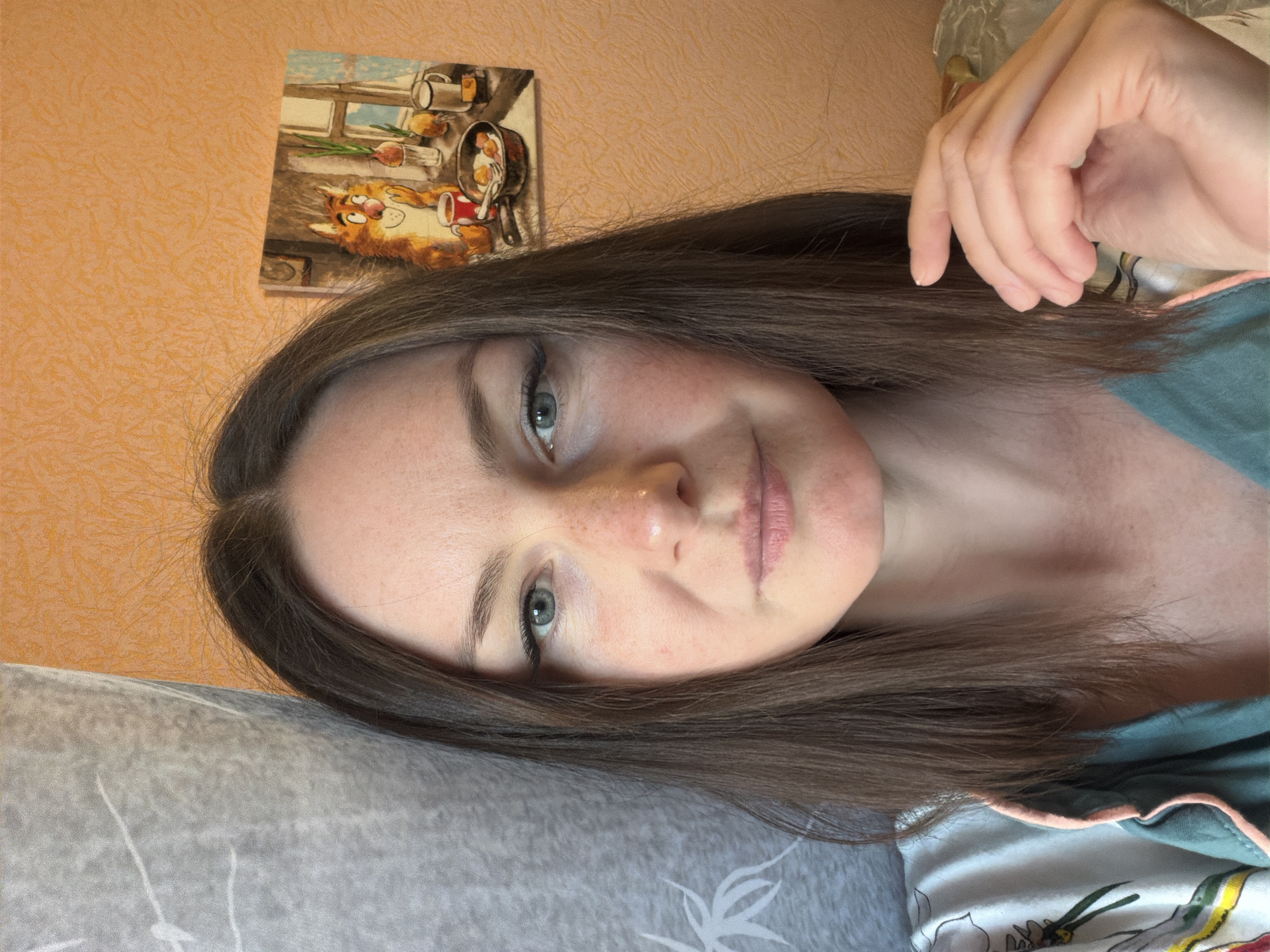 A close-up, slightly angled portrait shot depicts a woman with light skin, freckles, and light-colored eyes, likely taken indoors. Her dark hair is parted and falls around her face. She is resting her head on a gray pillow or cushion. In the background, an orange wall is visible, adorned with a framed picture of a cat in a kitchen scene. Part of her hand is visible in the top right corner of the frame. She is wearing a turquoise top with a pink-trimmed collar. The location is unknown.