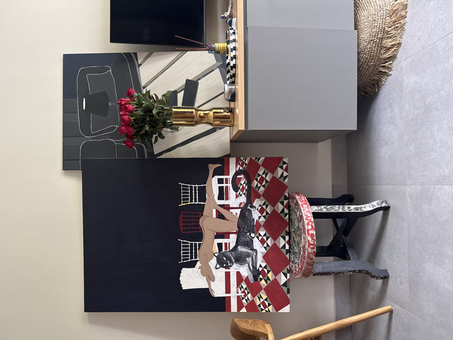 The image shows an interior space with various artistic elements. Three paintings hang on the wall. One depicts a minimalist chair, another showcases an upside-down nude figure with a black cat against a red and white patterned backdrop, and the third is an abstract linear design. A golden vase with red roses sits on a wooden surface. Below, there is a rustic wooden stool with a decorated round seat. A woven straw basket rests on top of a gray cabinet. A wooden broom leans against the wall.