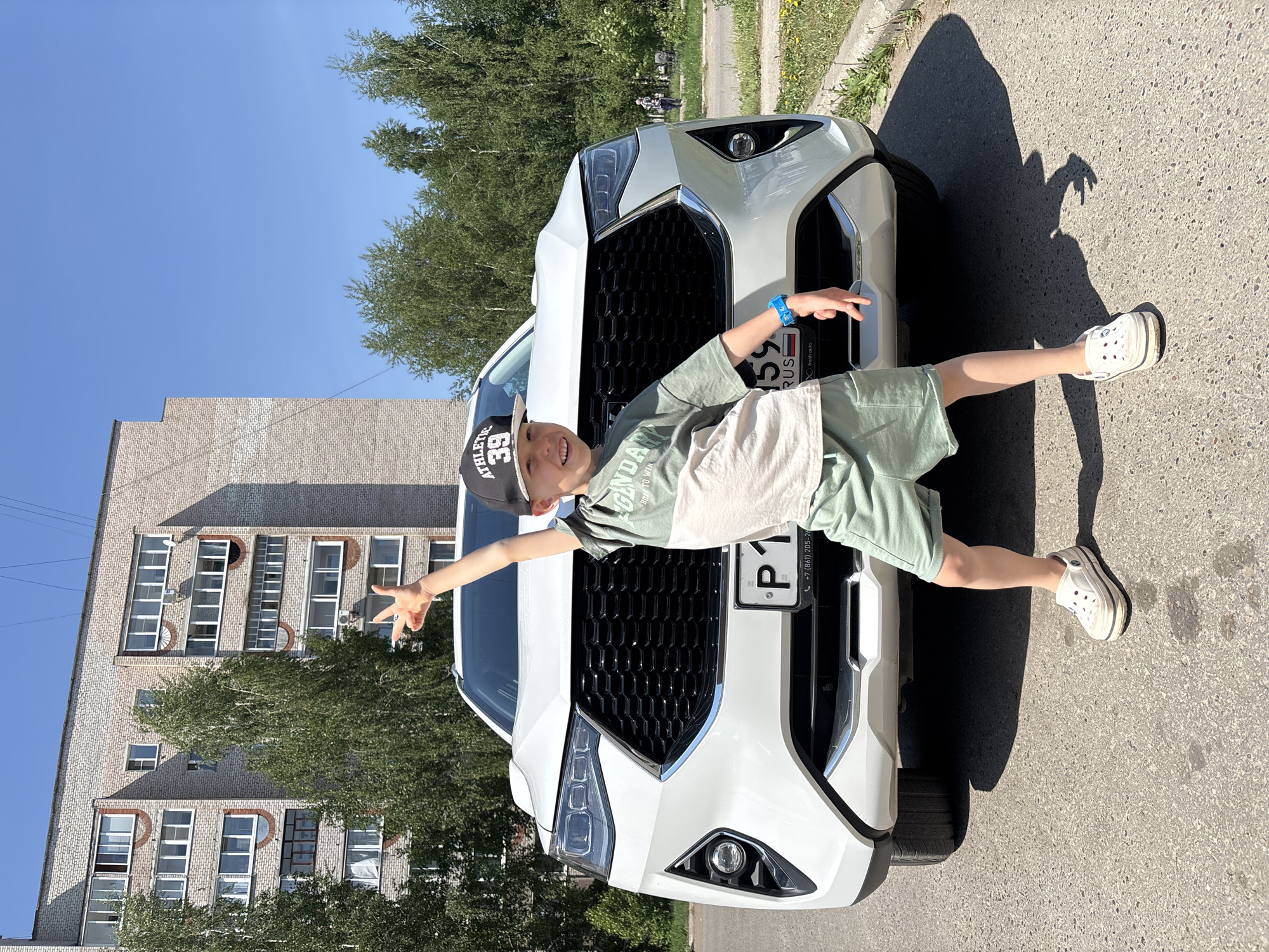 The image captures a young boy posing playfully on the front of a white car. The boy is wearing a baseball cap with the number "39" on it, a green t-shirt with a white patch, green shorts, and white sandals. He is smiling and making a peace sign with one hand while pointing with the other.
The car appears to be parked on a street near a building constructed from pale brown bricks with rows of windows. In the background, there are trees and a clear blue sky.
The license plate on the car reads "P159" and indicates a Russian origin. The image includes a sticker indicating "fresh auto." A readable phone number is visible on the license plate frame: "+7 (861) 205-26." Based on the license plate information and previous context, the location is likely Chaikovsky, Russia.