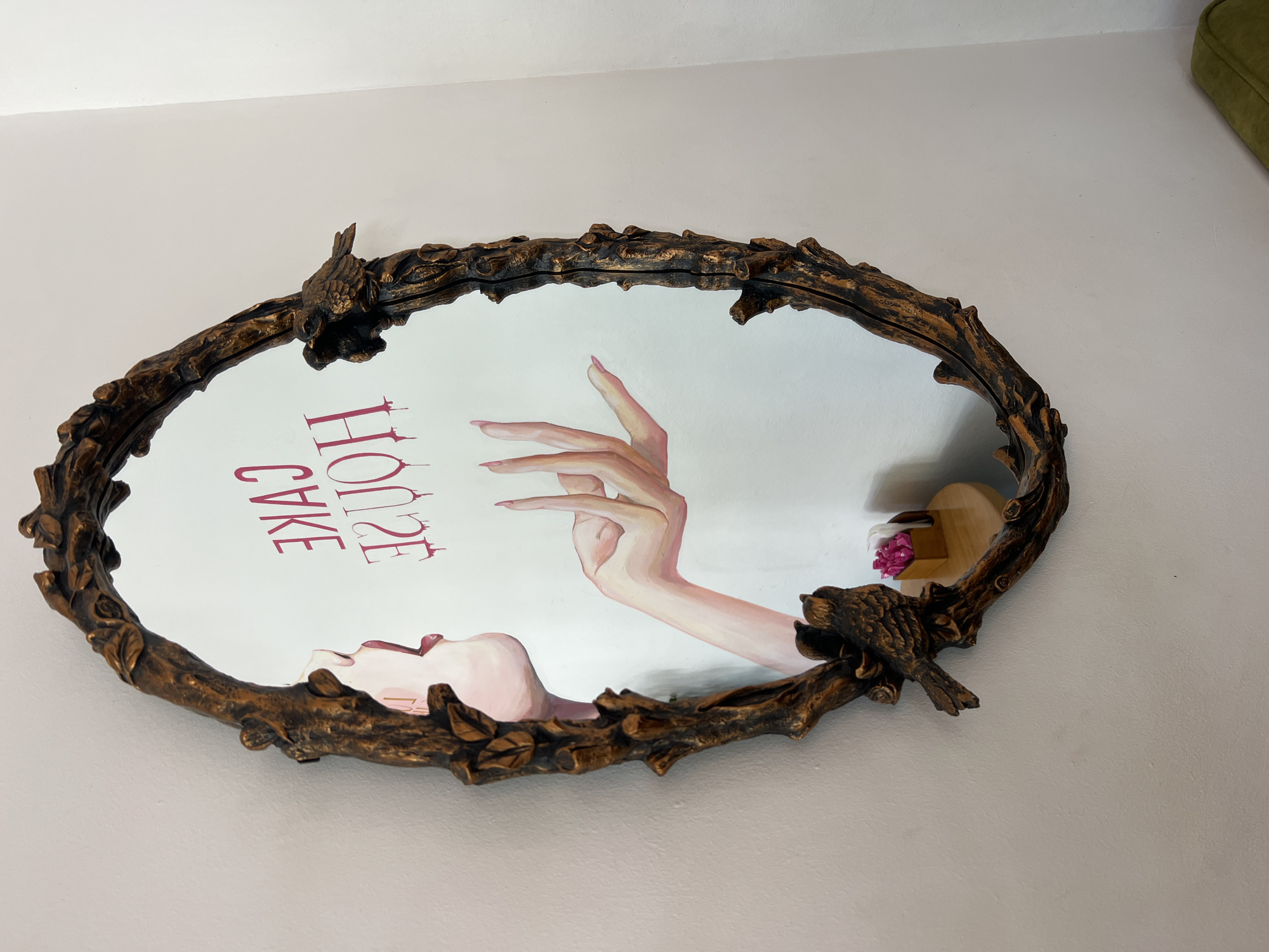 A highly ornate, oval mirror with a gold-colored, distressed frame resembling tree branches and adorned with two small bird figurines hangs on a light-colored wall. The mirror reflects an artwork featuring a stylized pink hand with long nails reaching upwards, and the words "CAKE HOUSE" in large, pink, dripping letters. The bottom right corner of the mirror shows a small wooden table with a napkin holder containing pink napkins. The scene is likely an interior, possibly a cafe or shop, given the text and the decorative elements. The location is Odesa, Ukraine.