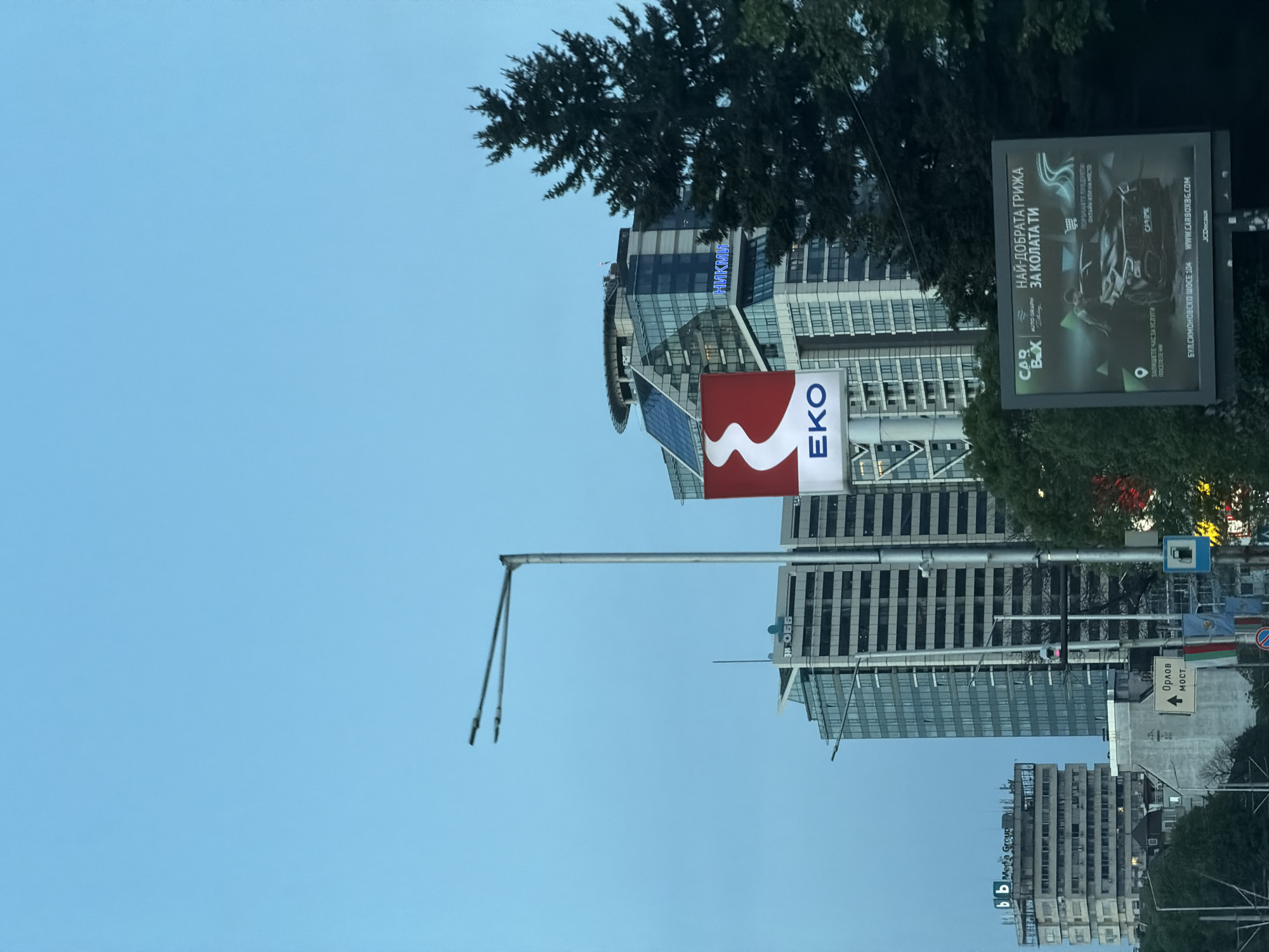 This image captures a modern cityscape in Sofia, Bulgaria, under a clear, light blue sky, likely during the day. The scene is dominated by several tall, contemporary buildings with glass and concrete architecture.
A large, prominent red and white sign with the "EKO" logo is displayed on the side of one building in the mid-ground. Above it, on a taller structure, blue illuminated text spells out "НИКИМ". On a building below the EKO sign, "ОББ" is visible, and on a lower building to the right, "bb Media Group" can be seen.
A large billboard stands prominently on the right side of the image, advertising "CAR BOX" with a graphic of a car. The Cyrillic text on the billboard reads: "НАЙ-ДОБРАТА ГРИЖА ЗА КОЛАТА ТИ" (The best care for your car). Additional text includes calls to action such as "ПОРЪЧАЙТЕ ПРОДУКТИ ОНЛАЙН ИЛИ НА МЯСТО" (Order products online or on-site) and "ЗАПИШЕТЕ ЧАС ЗА УСЛУГИ ПОСЕТЕТЕ НИ" (Book an appointment for services, visit us). A website, "WWW.CARBOXBG.COM", and an address, "БУЛ. СИМОНОВСКО ШОСЕ 104" (Bul. Simonovsko Shose 104), are also displayed, along with "JCDecaux" branding.
In the lower right, beneath the billboard, a white road sign points right with the text "Орлов МОСТ" (Orlov Most), a significant landmark in Sofia, confirming the geographical context. A tall metal street light pole with two fixtures extends into the sky on the left. Dark green tree foliage frames parts of the image, contrasting with the urban structures.