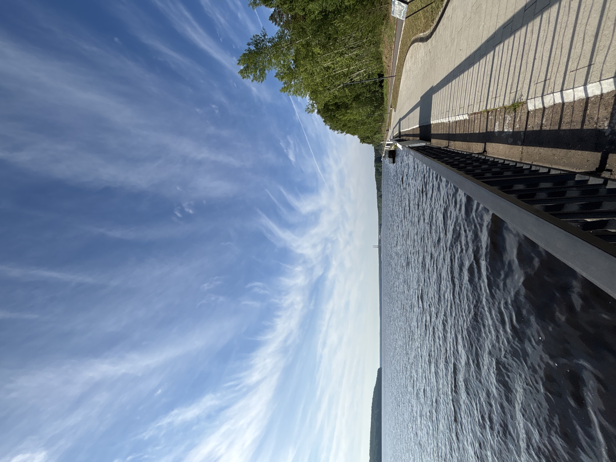The image presents a serene waterside scene under a bright, sunny sky. A vast expanse of calm water, possibly a river or lake, fills the lower portion of the frame, its surface textured with small ripples. Along the water's edge, a dark metal railing runs horizontally, separating the water from a paved promenade or walkway. Long, distinct shadows cast by the railing stretch across the light-colored paving, indicating a bright sun, likely during mid-morning or late afternoon.
Beyond the walkway, a dense line of green deciduous trees with lush foliage forms a natural barrier, suggesting a park or forested embankment. A small, white sign with indistinct black text is partially visible among these trees on the left side of the image. The sky above is a clear, vibrant blue, adorned with delicate, wispy white clouds, possibly cirrus or cirrocumulus, and a faint contrail from an aircraft is also visible. Across the water, a low-lying landmass is faintly discernible on the distant horizon. No people or specific activities are visible, contributing to the tranquil atmosphere. The setting is Chaikovsky, Russia.
