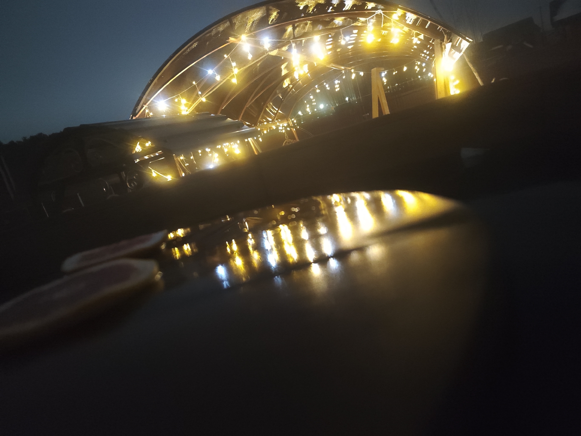 The image depicts a dimly lit outdoor scene, likely at night, in Chaikovsky, Russia. The primary focus is a low-angle view of a surface reflecting numerous illuminated lights.
Beyond the surface, the lights appear to originate from a structure resembling a tunnel or a large archway adorned with strings of decorative lights. The structure seems to be constructed of wood or similar material. The lights are predominantly yellow or warm white in color, suggesting incandescent bulbs or LEDs.
The scene's darkness obscures details, but there appear to be objects such as a table or bench behind the surface on which lights reflect. In the extreme foreground, there are a few red objects.
Overall, the image captures a nighttime scene with a focal point on illuminated lights reflected on a surface, adding a decorative glow to the setting.