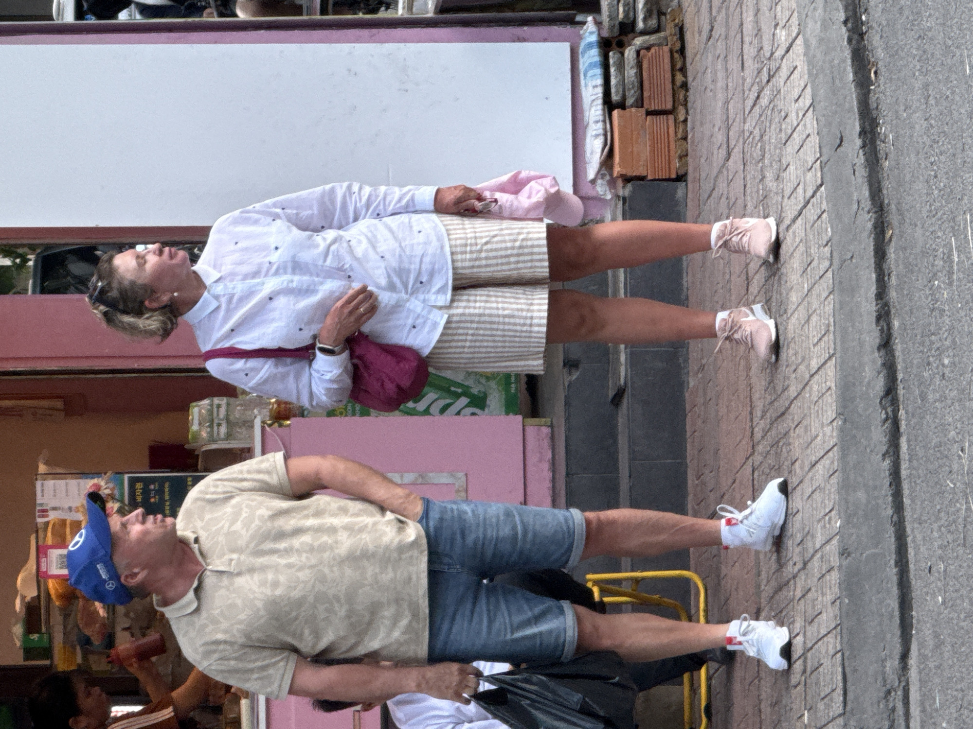 Two individuals, a male and a female, stand on a sidewalk in what appears to be a commercial area in Nha Trang Ward, Vietnam. The male, wearing a light-colored patterned polo shirt, denim shorts, and white sneakers, stands with his left hand in his pocket. He also wears a blue baseball cap. To his right, the female is dressed in a white collared shirt with dark patterns and striped shorts, paired with pink sneakers. She carries a pink bag over her left shoulder and holds a pink folded item in her right hand. Behind them, a pink-fronted building suggests a shop or restaurant. The sidewalk is paved with stone tiles, and a section of asphalt road is visible in the foreground. Construction materials, including bricks and a striped mat, are stacked to the right of the scene. The environment appears to be outdoors during daylight.