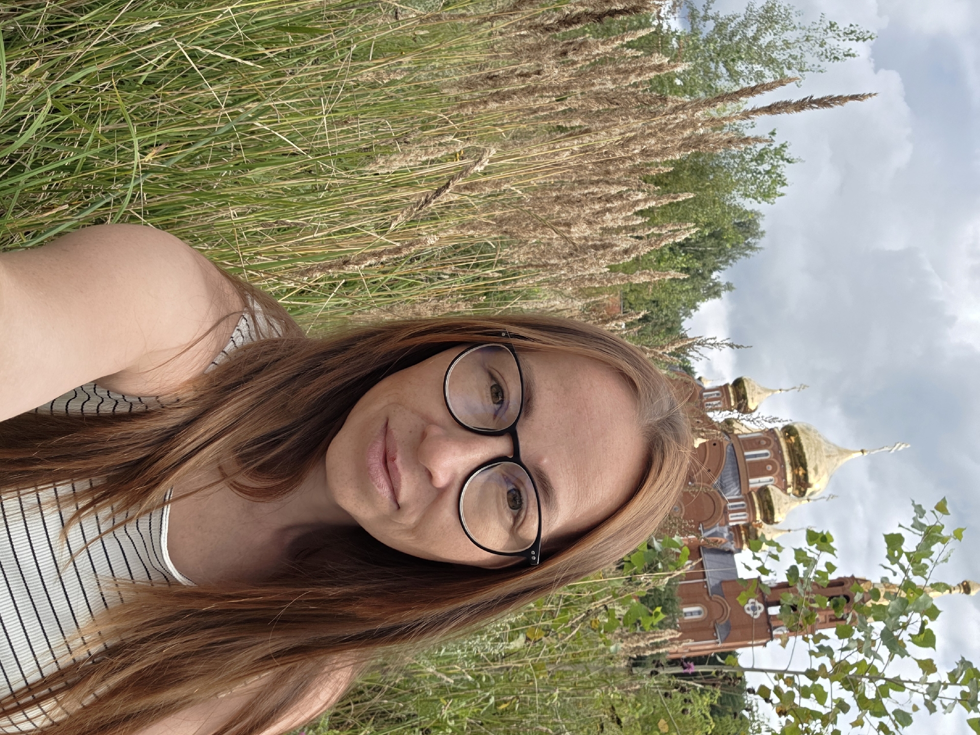 A woman with long, reddish-brown hair and glasses is taking a selfie in front of a Russian Orthodox church with golden domes. She is wearing a striped tank top. The church is made of red brick. Tall, dry grass and green foliage surround the woman and the church. The sky is overcast with clouds. The location is the Holy Trinity Cathedral in Chaikovsky, Russia.