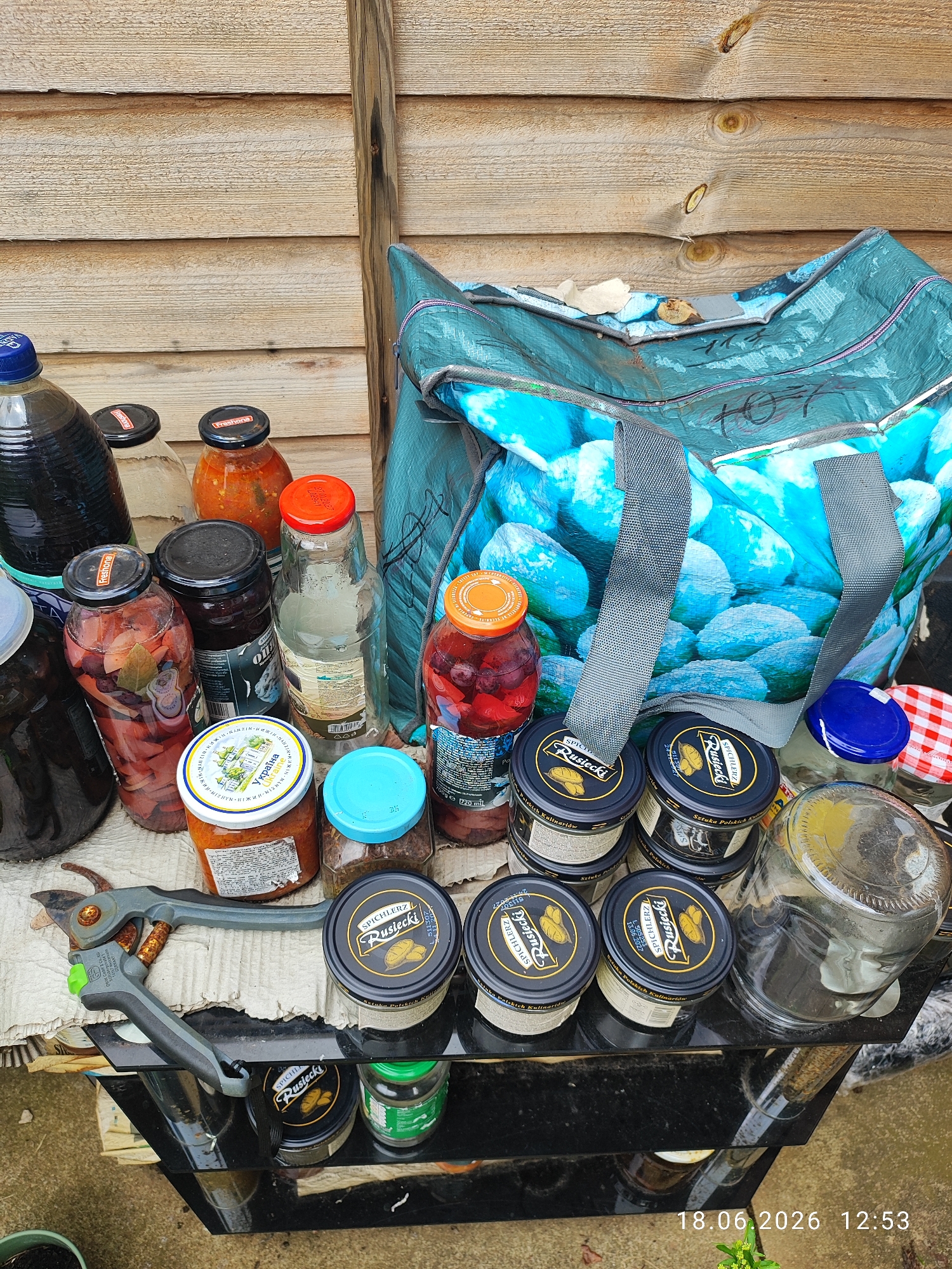 This is a high-angle, eye-level shot of a collection of jars, bottles, and a reusable shopping bag filled with what appears to be preserves or pickled items, arranged on a rough surface, possibly a tarp or old fabric, with some items placed on a black metal shelf. The scene is outdoors, with a weathered wooden fence in the background, suggesting a garden or backyard setting. The lighting indicates it's daytime, and the overall atmosphere is casual and utilitarian, possibly a collection of homemade goods or items being stored.
Prominently featured are several glass jars containing various colored preserved fruits and vegetables, some with visible spices like bay leaves. There are also tins of "Spichlerz Rusiecki" Polish culinary items. A large blue and white patterned reusable shopping bag, possibly containing more of the same, dominates the right side of the frame. A pair of gardening shears lies in the foreground, hinting at activities related to harvesting or preserving produce.
Visible text includes brand names like "Freshona" and "Spichlerz Rusiecki," along with some Ukrainian text on one of the jar labels. A timestamp "18.06.2026 12:53" is present in the bottom right corner. The scene does not contain any people, but the presence of the gardening shears suggests a person has recently been or will soon be involved in activities related to these items. The setting appears to be a domestic outdoor space, likely in Northampton, United Kingdom, given the provided context.