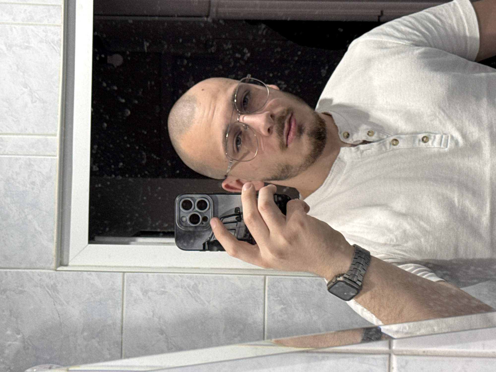 A male individual is captured in a mirror selfie, seen from the chest up in an indoor setting. He has a shaved head, a short beard and mustache, and wears wire-rimmed eyeglasses. He is dressed in a light-colored, short-sleeved Henley shirt featuring multiple brown buttons. A smartwatch with a metallic link band is present on his left wrist. The subject holds a dark-cased smartphone in his right hand, angled to photograph his reflection. The phone case displays a monochromatic graphic, suggestive of a vehicle. The background reveals light grey rectangular wall tiles with visible grout lines. A dark, speckled area, outlined by a white frame, is visible in the reflection behind the subject. The image contains no visual elements that allow for the identification of the location as Stara Zagora, Bulgaria, or any specific urban area.