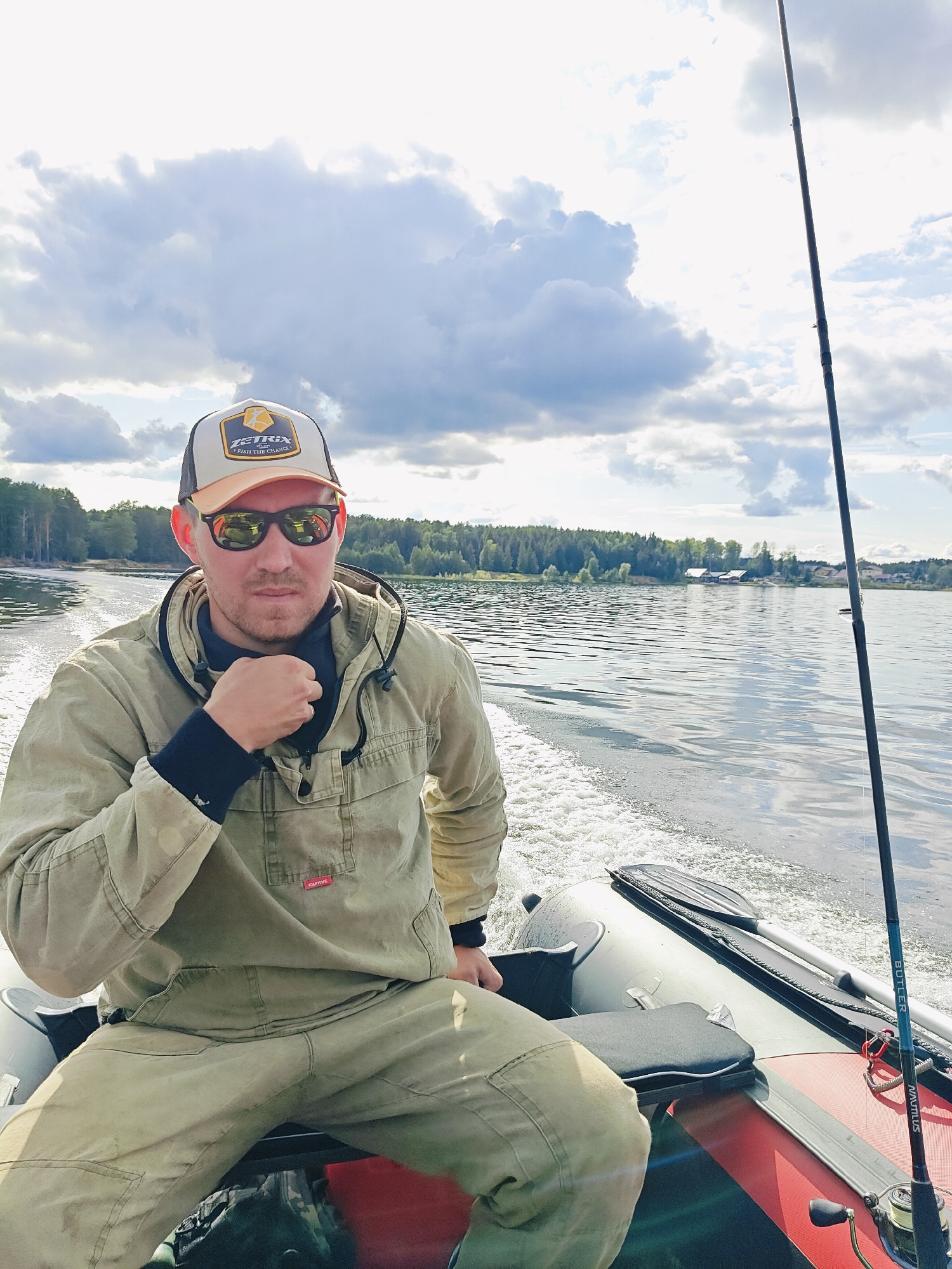 A man is sitting in an inflatable boat on a body of water, possibly the Kama River, near the city of Chaykovsky, Russia. The man is wearing a tan fishing suit, a baseball cap, and sunglasses. He is holding his hand to his chin and looking at the camera. A fishing rod is visible in the boat next to him. The water is calm, and there are trees and buildings on the shore in the background. The sky is cloudy.