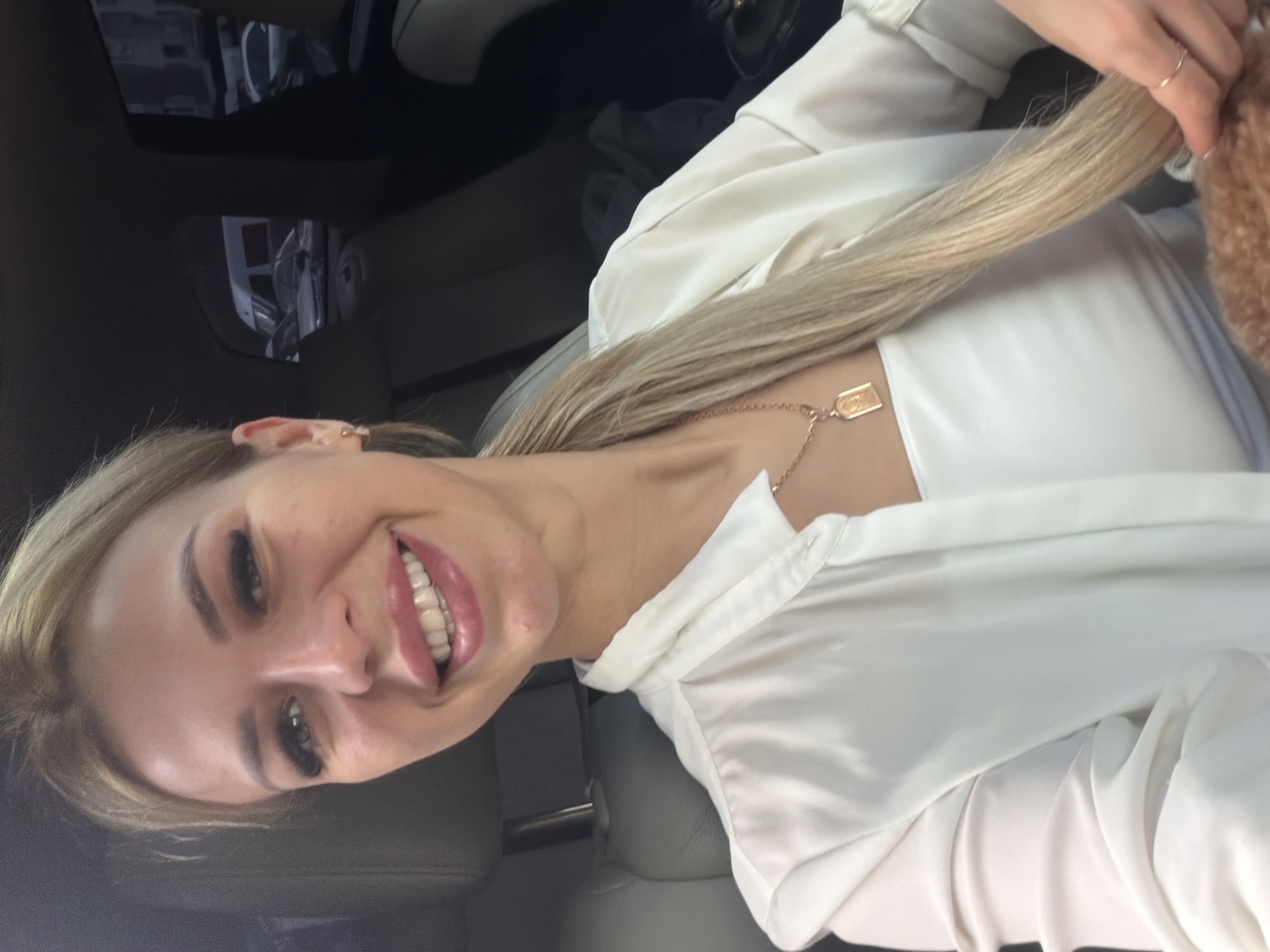A blonde woman with light skin and dark eyebrows is sitting in the passenger seat of a car, smiling at the camera. She is wearing a white collared shirt over a white top, and a gold pendant necklace. Her blonde hair is pulled back into a ponytail that falls over her shoulder. The interior of the car, including grey leather seats, is visible in the background. Through the windows, other cars and buildings can be seen, suggesting an urban environment. The woman is touching her hair with her right hand, wearing a ring on her ring finger.