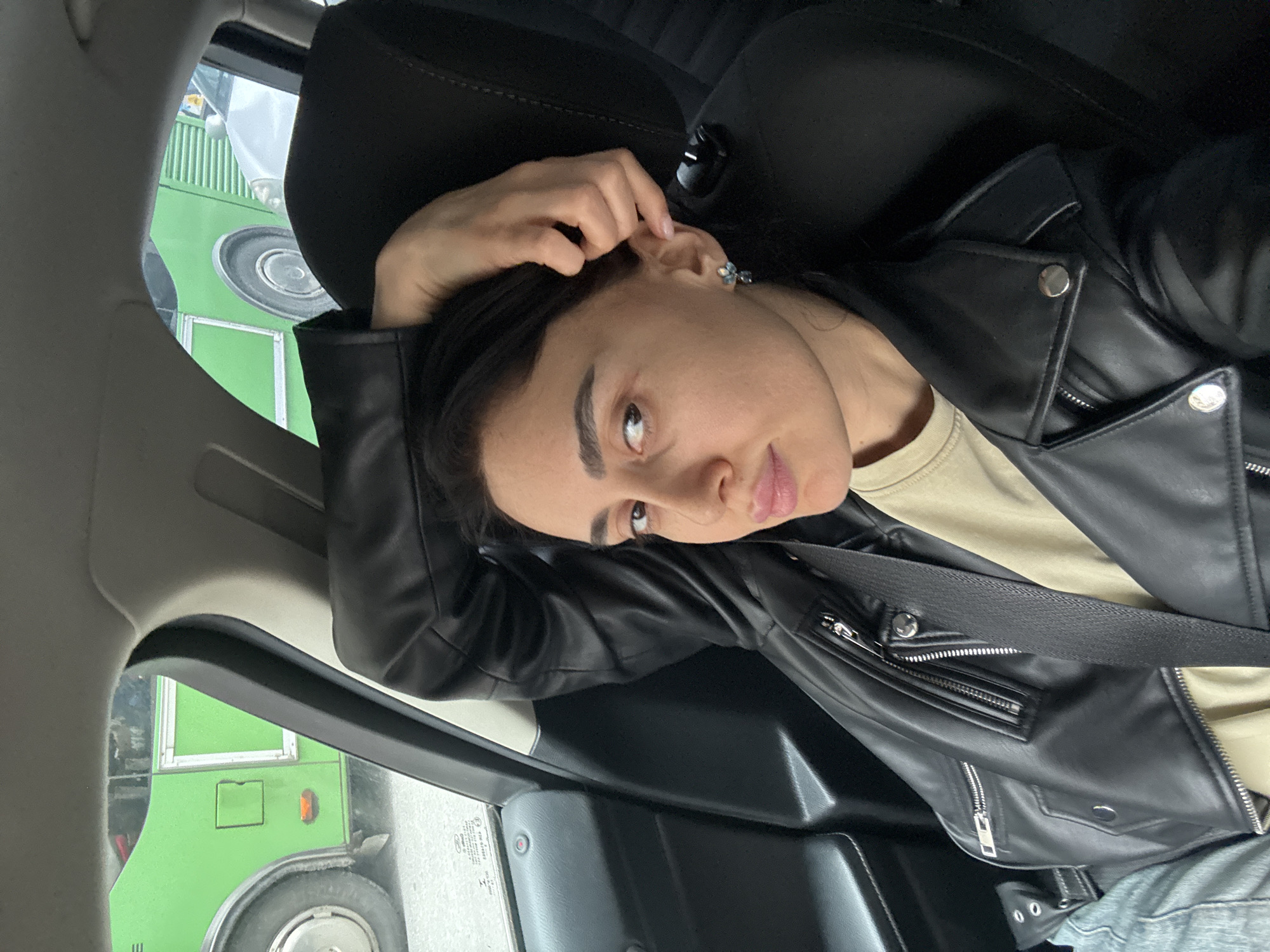 A female individual is positioned in the front passenger seat of a vehicle, facing the camera. She has dark hair and a light-to-medium complexion. She is dressed in a black faux leather jacket with silver zipper and snap detailing, worn over a light beige crew-neck shirt, and blue denim trousers. A black seatbelt is diagonally secured across her torso. Her left arm is flexed, with the hand resting on the back of her head, fingers positioned near her right ear. A multi-petal, light-colored earring is affixed to her right earlobe. The vehicle's interior is dark, with a black headrest and door panel visible. Visible through the front passenger window is the side profile of a bright green public transport bus, featuring a wheel, a lower panel, and a portion of a side window. A second, lighter-colored vehicle is partially visible behind the bus. The characteristic green public buses identify the scene as occurring within the city of Tbilisi, Georgia.