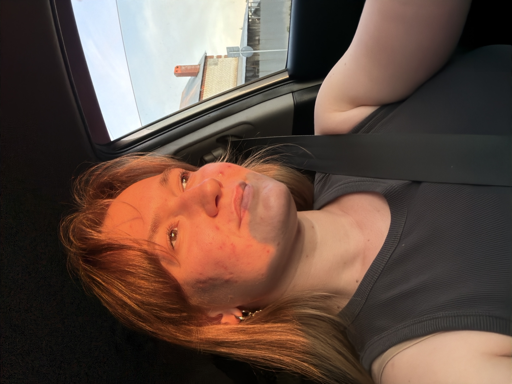 A young woman with blonde hair and fair skin is pictured inside a car, looking out the window to her right. She is wearing a dark gray tank top and a seatbelt. The setting sun casts a warm, golden light on her face, highlighting some skin imperfections. Outside the car window, a brick building with a red-tiled roof and a fence are visible under a pale sky. The image appears to be taken in Novokuybyshevsk, Russia, based on the context provided.