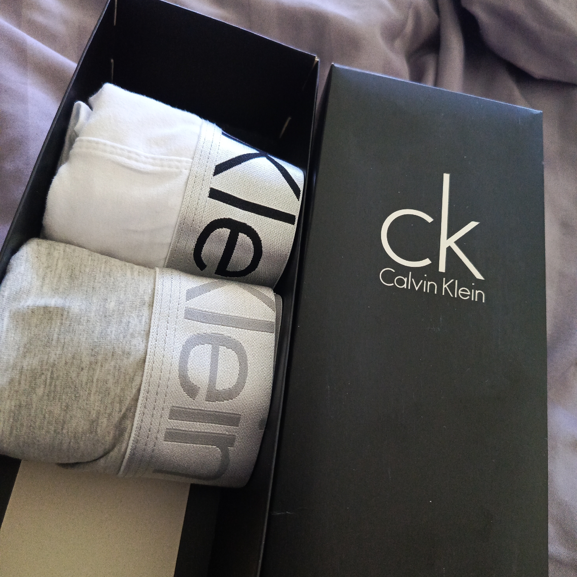The image shows a black Calvin Klein box containing two pairs of underwear. One pair is white, and the other is gray. The box is sitting on a lavender-colored surface, possibly a bedspread or blanket. No people, subjects, actions, or interactions are visible. The location is not evident in the image.