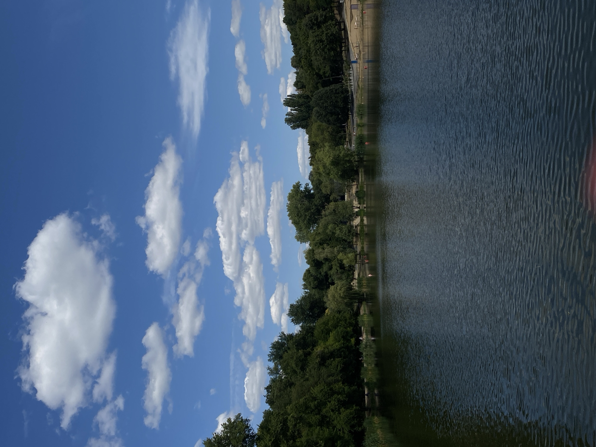 The image depicts a scenic view of a lake on a clear day with scattered white clouds in a bright blue sky. The foreground is dominated by the dark, rippling surface of the lake. Along the far bank of the lake, a dense line of lush green trees forms a natural boundary, with varying shades of green indicating different types of foliage and heights.
In the background, beyond the trees, is a sandy beach area. Several small red buoys are visible in the water near the beach. A few individuals are present on the beach and by the water's edge, appearing to be relaxing or engaged in recreational activities. The overall impression is of a tranquil park or recreational area within an urban setting.
This scene is characteristic of the park and lake complex located in Chișinău, Moldova, specifically the Valea Morilor Park (also known as the "Mill Valley Park"). This park is a popular spot for recreation and relaxation, featuring a large artificial lake, walking paths, and green spaces.