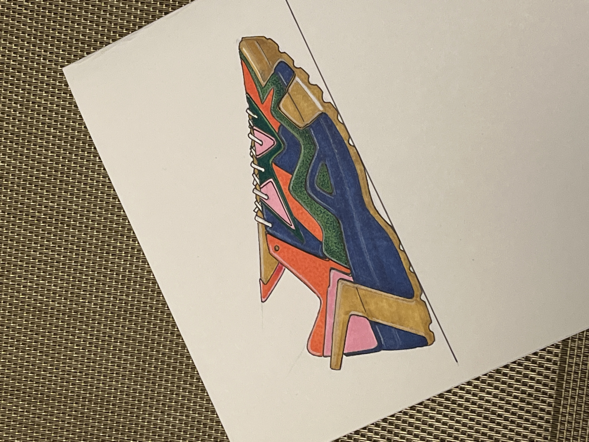 A drawing of a geometric shoe on white paper is positioned on a brown woven surface. The shoe is rendered in various colors, including gold, blue, green, orange, and pink. It has a jagged sole and laces. The paper is angled on the woven surface, with a thin black line running down its right side. The drawing is not located in any city or location.