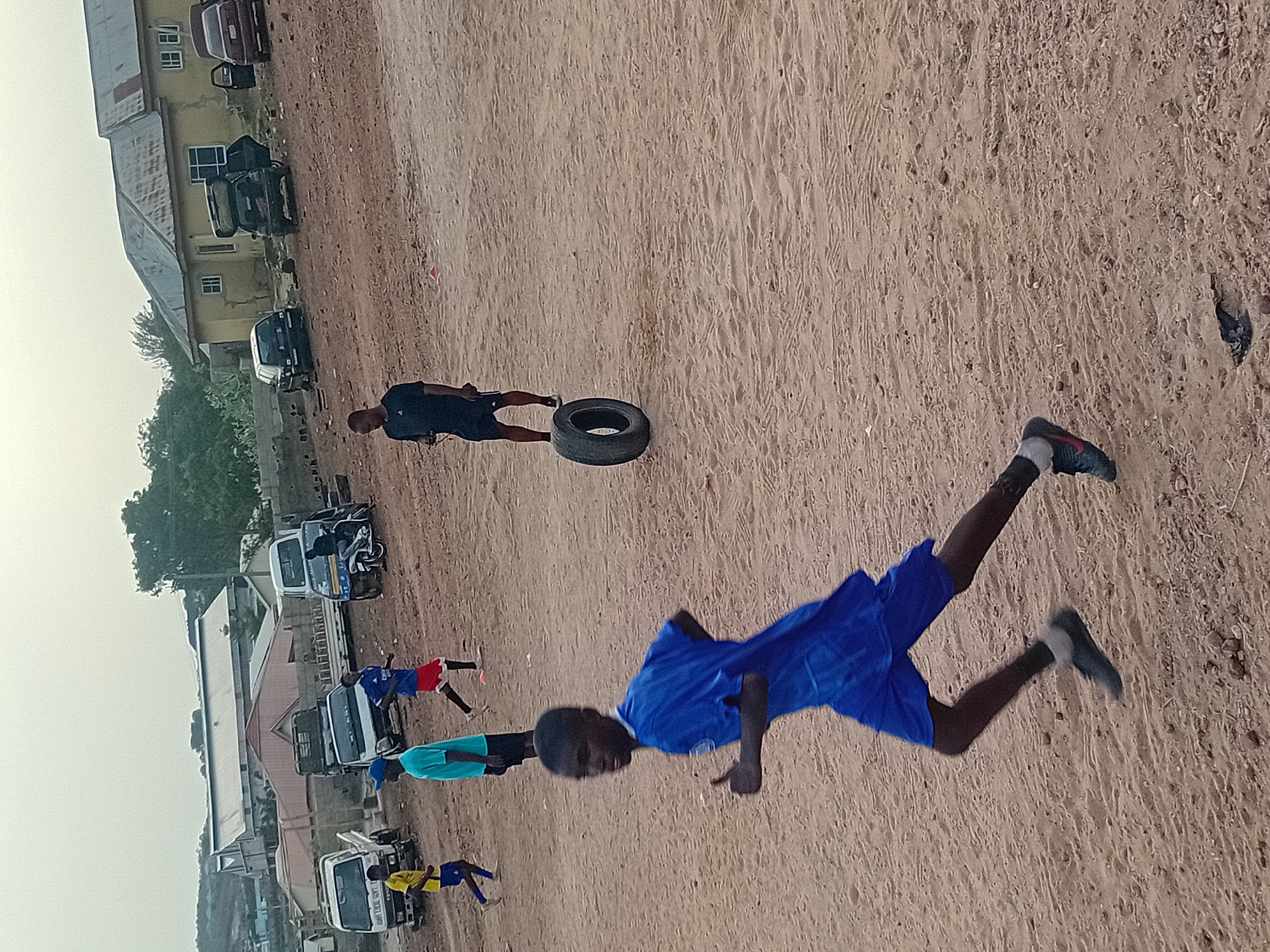 An outdoor scene depicts several individuals, likely young males, engaged in what appears to be an informal athletic training session or sports activity on a reddish-brown dirt field.
In the foreground, slightly to the right, a person with dark skin, wearing a royal blue t-shirt and shorts, is captured mid-action, possibly jumping or running, with their arms bent and legs extended. They wear dark sports shoes with white accents. Further back and to the left, another person with dark skin, dressed in a dark t-shirt and dark shorts, stands next to a discarded black car tire, suggesting it might be used for exercise.
In the mid-ground and background, additional individuals are visible. One person wears a blue and red outfit, another a light blue/teal t-shirt with dark shorts, and a third is in a yellow and blue uniform, partially obscured by a white truck.
The setting includes a collection of buildings in the background, varying in color from yellowish to white/grey, some with visible windows. Several vehicles, including various white trucks, a dark SUV, and a burgundy car with an open door, are parked along the edge of the dirt area. Some faint text like "TOYOTA" can be seen on the sides of a few trucks. Green trees and other vegetation are visible behind the buildings on the left.
The ground is unpaved, uneven, and dusty, with small rocks and footprints. The sky is light and hazy, suggesting an overcast day or possibly early morning/late afternoon lighting. The overall impression is one of communal outdoor physical activity in a semi-urban or rural environment.