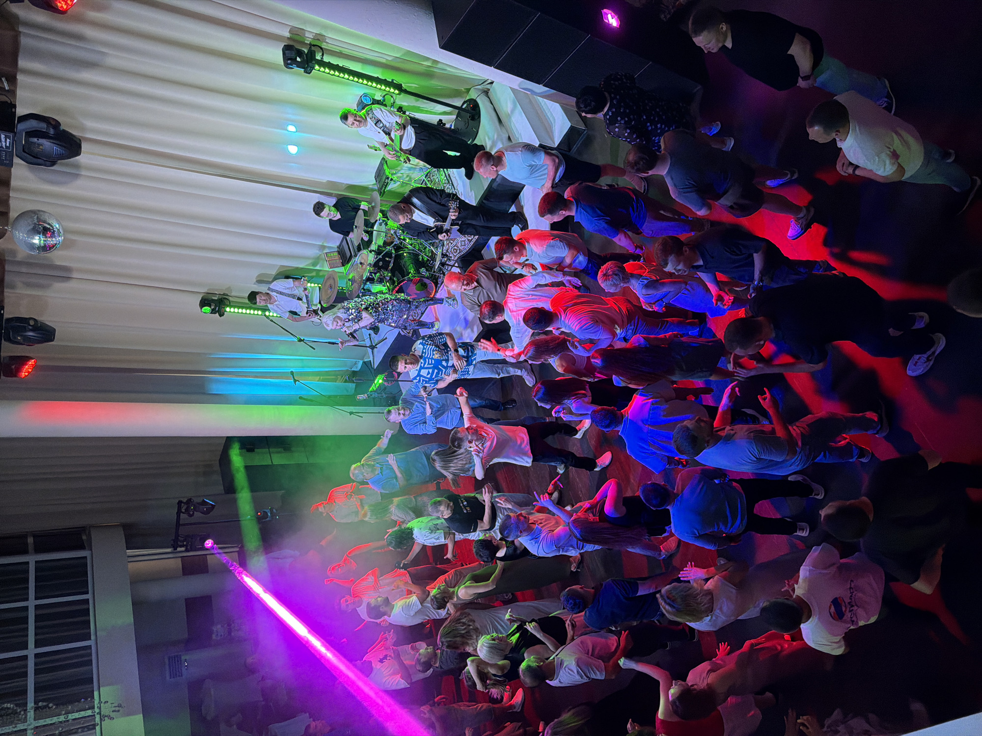 The image captures a lively indoor event, likely a concert or a party, taking place in Chaikovsky, Russia. The scene is dominated by a crowd of people, mostly adults, gathered to watch a band performing on a stage. The band members, illuminated by colorful stage lights, are actively engaged in their music. One guitarist is visible in the foreground, wearing a white shirt and playing his instrument, while another member is behind a drum set. The audience is a mix of men and women, some standing and observing, while others appear to be dancing or enjoying the performance.
The setting is an indoor venue with a stage adorned with bright green and pink lights, creating a vibrant and energetic atmosphere. A disco ball is also visible, adding to the festive mood. The background features large, dark speakers, typical of a performance space. The lighting is dynamic, with beams of purple and green light casting dramatic shadows and reflections on the floor and the people. The overall mood is one of excitement and shared enjoyment.
There are no discernible weather cues as the event is indoors. The time of day is difficult to determine precisely, but the lighting suggests it's likely evening or night. The location is confirmed to be Chaikovsky, Russia. There is no visible text in the image.