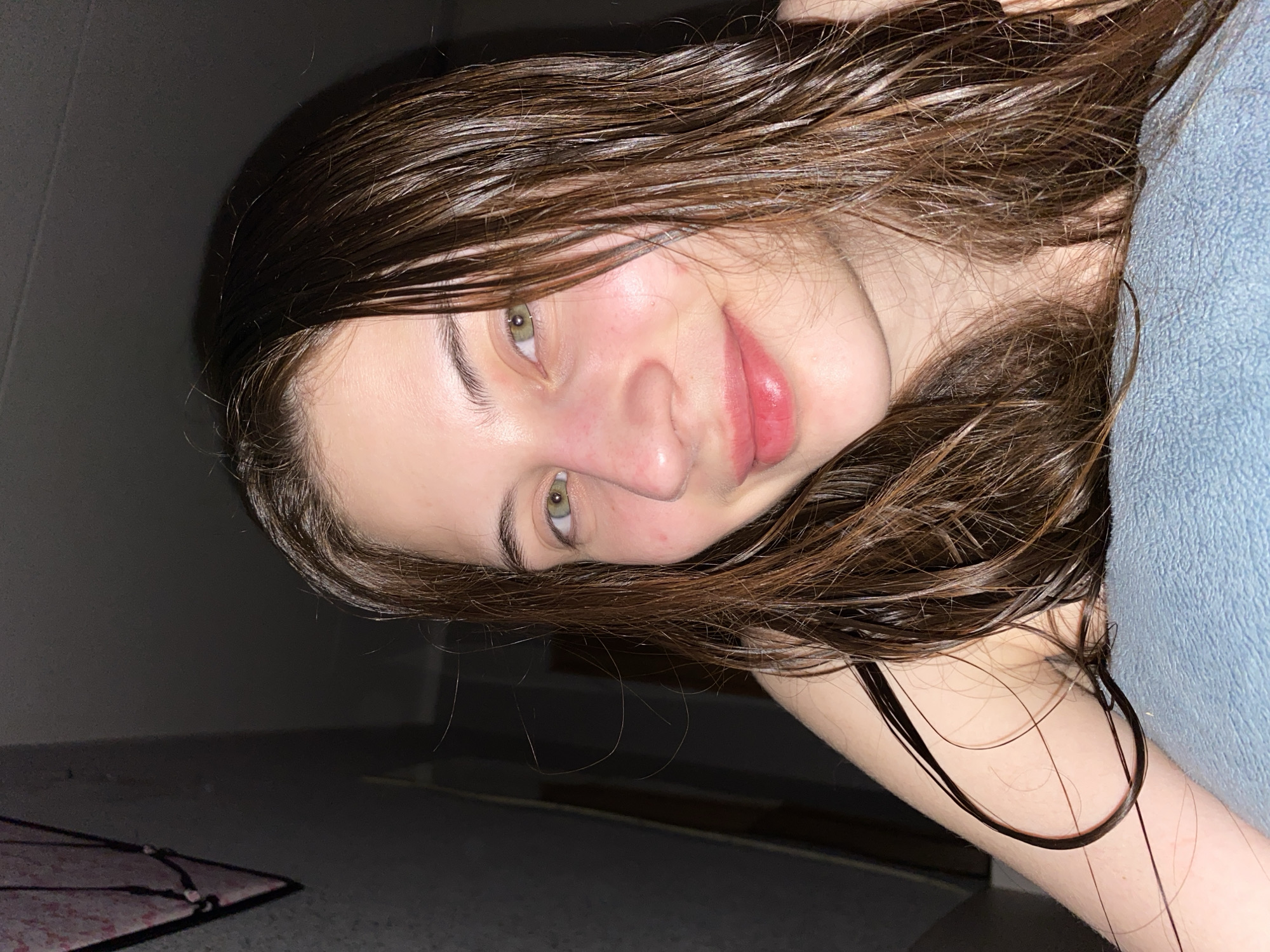 A young woman with long, dark brown hair and striking green eyes is shown in a tilted, close-up portrait, presumably taken in Votkinsk, Russia. Her hair appears wet or freshly washed, with individual strands visible, and cascades over her shoulder and arm. She is lying down, resting her head on a blue, towel-like fabric, part of which is visible on the right. Her bare arm is also visible in the lower right. Her skin is clear, with a natural flush on her cheeks, and her lips are a soft pink. She looks directly at the camera with a calm, slightly smiling expression. The background is dark and out of focus, indicating an indoor setting with low ambient light, likely evening or night. A faint, dark patterned object, possibly a mat or textile, is visible on the floor or surface in the lower left corner. The lighting highlights her face and the wet sheen of her hair. No visible text is present in the image.