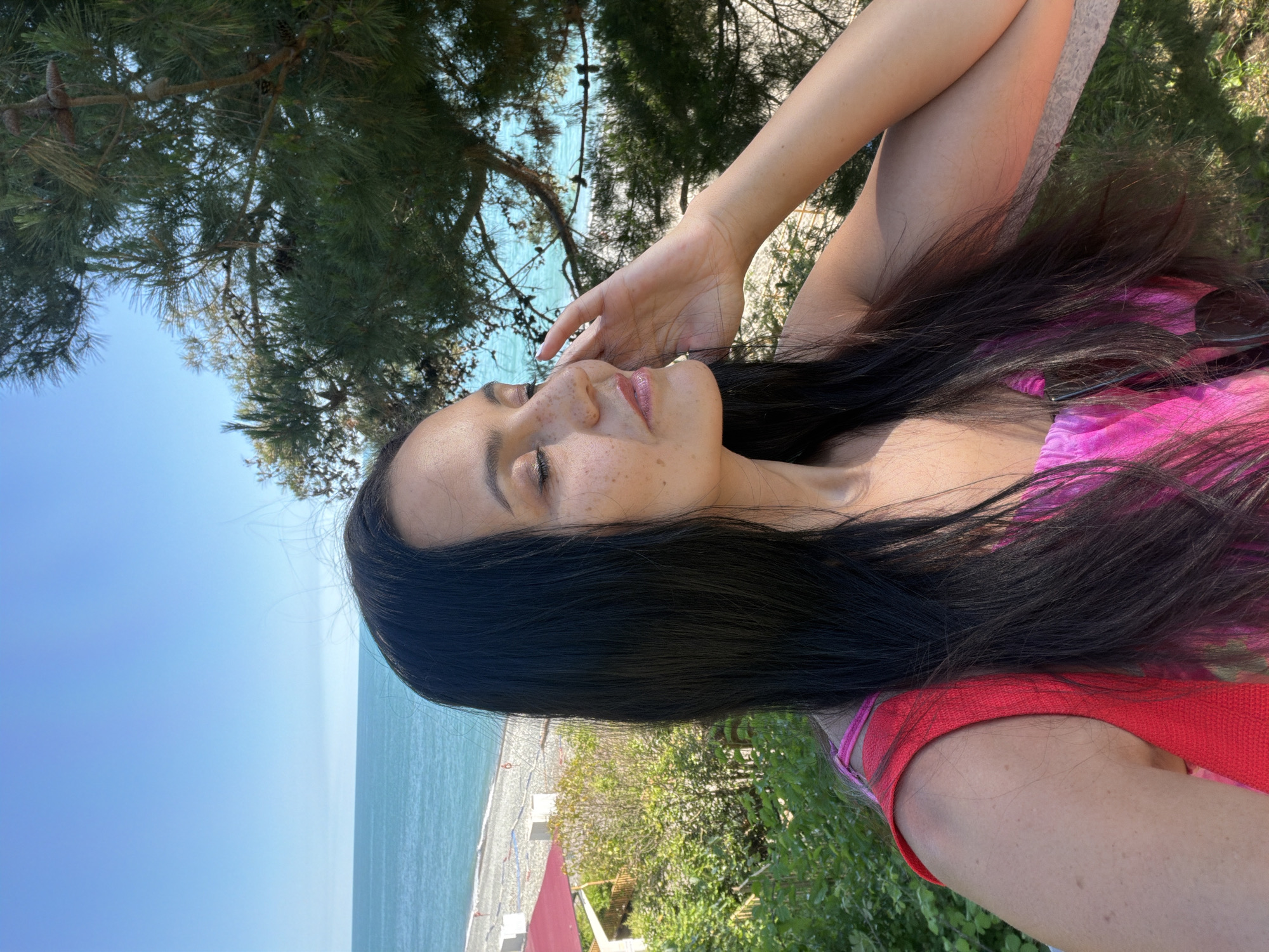 A profile shot of a woman with long, dark hair, freckles, and closed eyes is captured outdoors. She is wearing a vibrant pink and red dress and appears to be touching her face with her right hand. The background reveals a sunlit scene with a view of the turquoise sea and a pebble beach, characteristic of the coastal city of Sochi, Russia. Lush green foliage and a large pine tree are also visible. The overall atmosphere is peaceful and serene, suggesting a moment of relaxation by the sea.