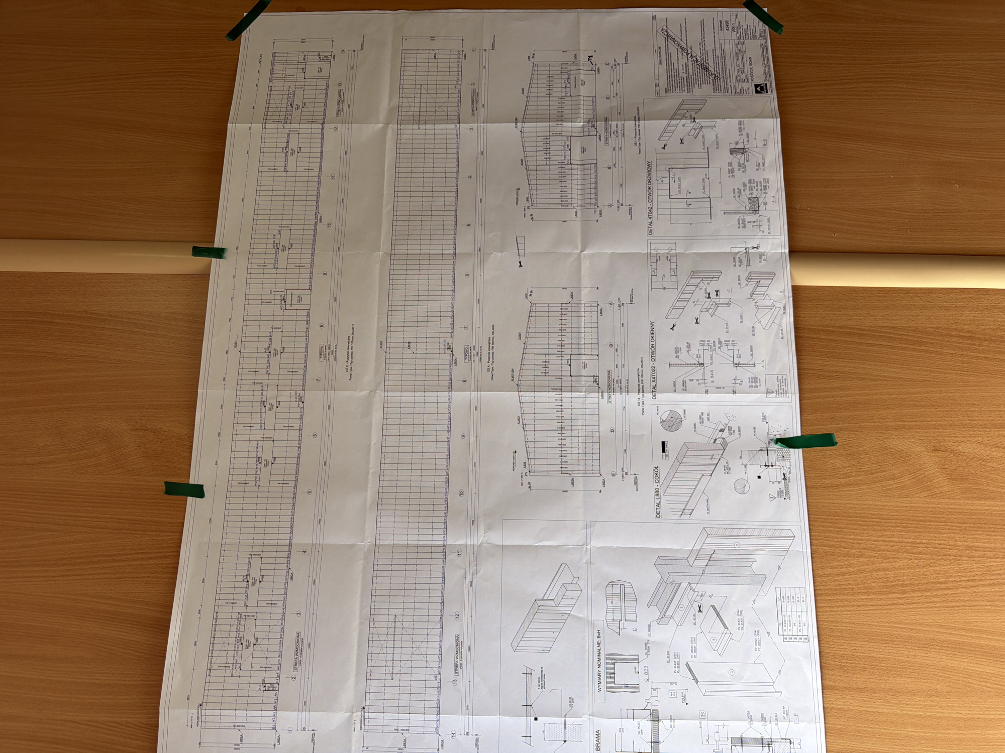 The image shows a large, unfurled architectural blueprint attached to a wooden wall with green tape. The blueprint is detailed, with grids, lines, text, and technical drawings. It appears to be a plan for a building, possibly a warehouse or industrial facility, as indicated by labels like "BRAMA" (gate) and "STREFA WZMOCNIONA" (reinforced zone). There are also sections labeled with numbers and possibly dimensions, along with various technical details and diagrams of building components. The overall impression is that of a construction project in progress, with the blueprint serving as a central document for planning and execution. The background is a plain wooden wall, suggesting an interior space, possibly an office or a temporary site office. The lighting is natural, coming from an unseen source, casting a soft light on the blueprint and wall. There are no people visible in the image. The location in the city of Stróża, Poland is mentioned, but there is no visual information within the image itself that directly indicates this specific location. The image is a static representation of the blueprint and its immediate surroundings.
