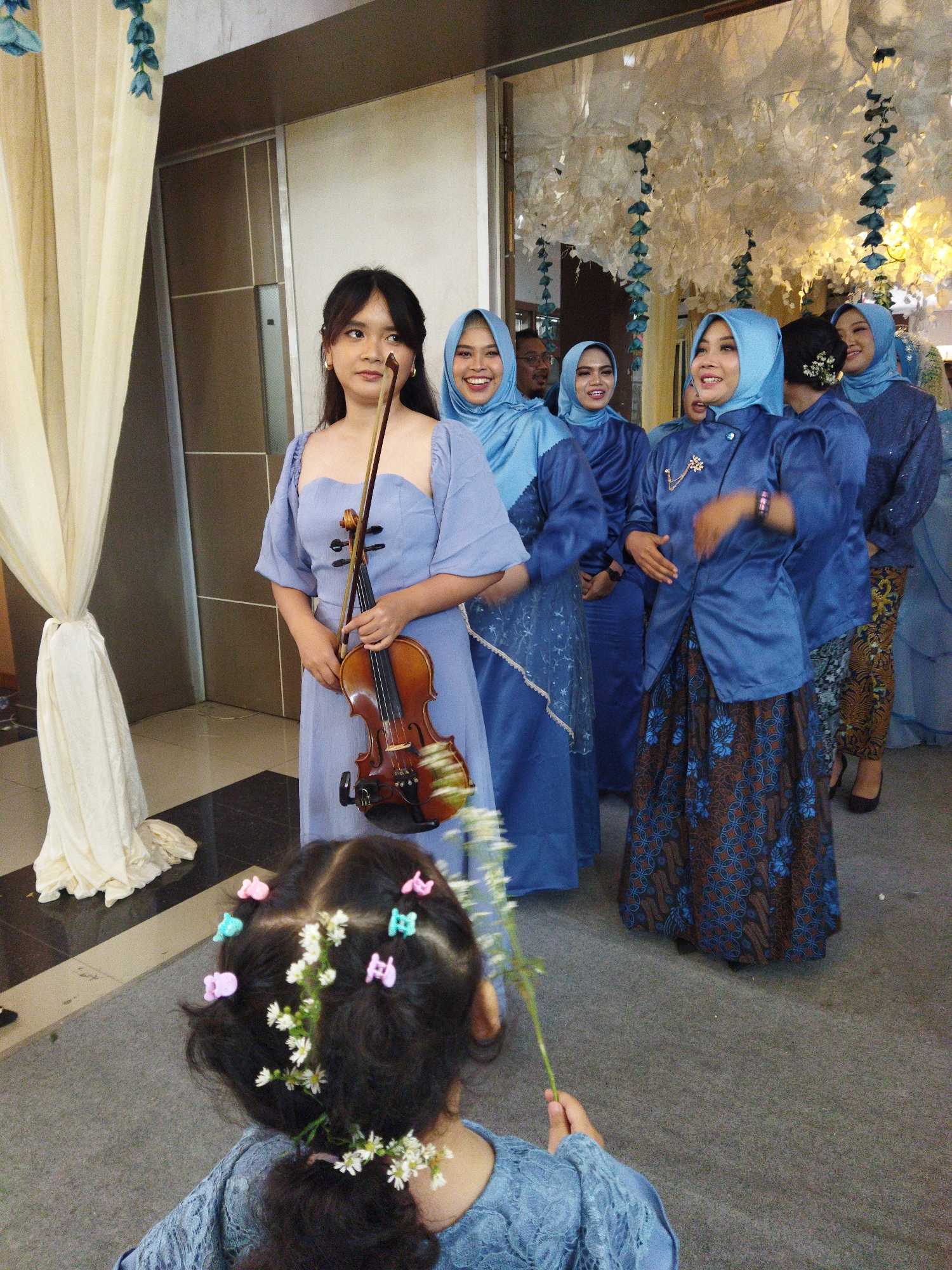 In a dimly lit indoor space, possibly a wedding reception or formal event venue in Semarang, Indonesia, a young woman stands center frame, holding a violin and bow. She wears a light blue, off-the-shoulder dress. Behind her, a group of women in matching blue hijabs and dresses are visible, some smiling and looking towards the camera. The woman on the far right is wearing a dark blue patterned skirt and a blue hijab, and the woman next to her is wearing a blue hijab and a blue top with a patterned skirt. A child in the foreground, with her back to the camera, has her hair decorated with small flowers and colorful hair clips. The environment features light-colored drapes on the left and decorative white and blue floral arrangements hanging from the ceiling in the background. The floor is carpeted in a neutral gray. The overall scene suggests a celebratory gathering.