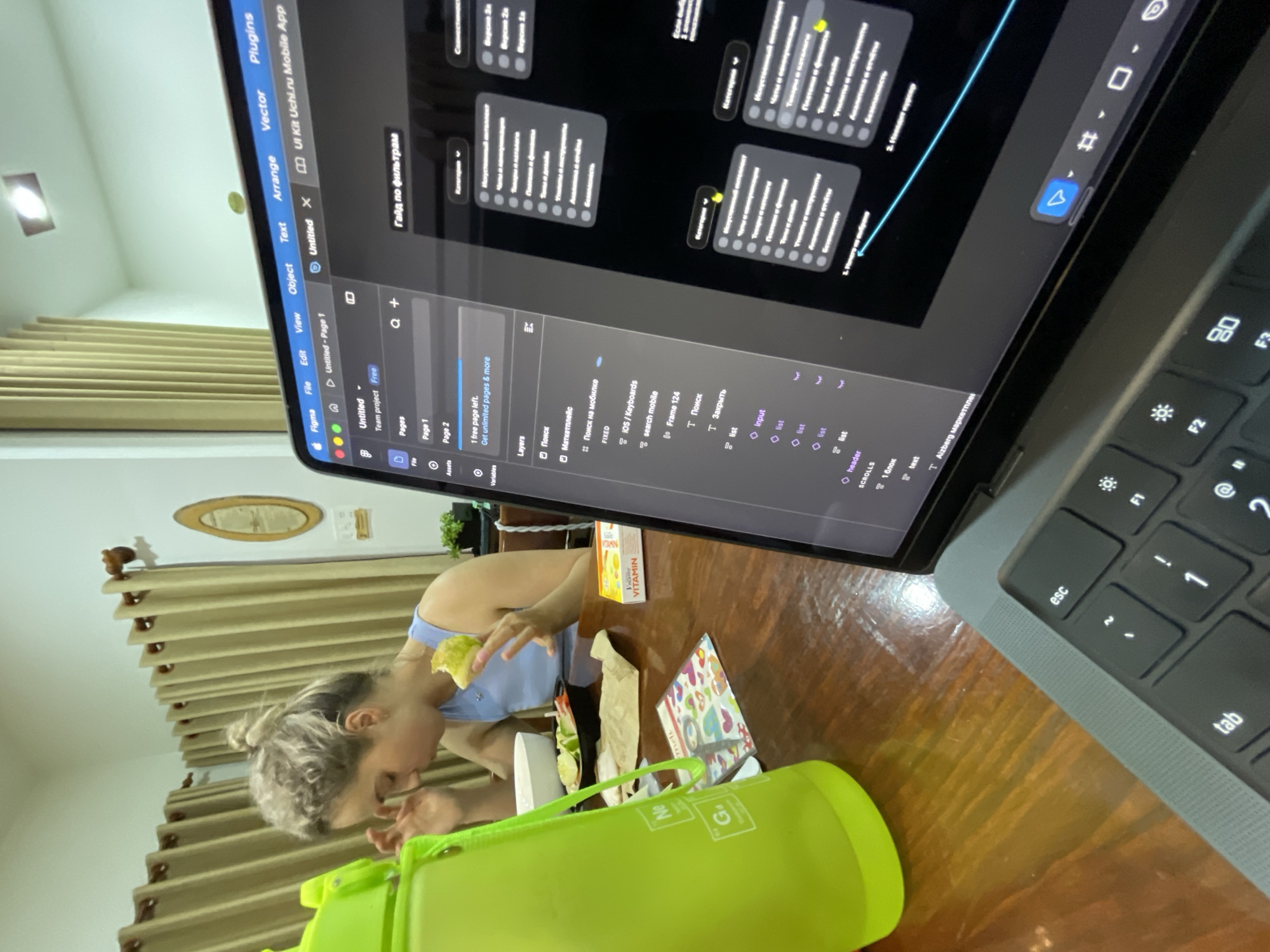 The image depicts a person with blonde hair eating at a wooden table in what appears to be a home setting in Lâm Viên - Đà Lạt, Vietnam. They are wearing a blue top and there is a green water bottle, a bowl, a sandwich, and what appears to be vitamins on the table. A laptop is open in front of them, displaying a design or development interface, possibly Figma, with UI elements and text in Russian. The laptop keyboard is visible to the right. The background includes curtains and a decorative oval plaque on the wall.