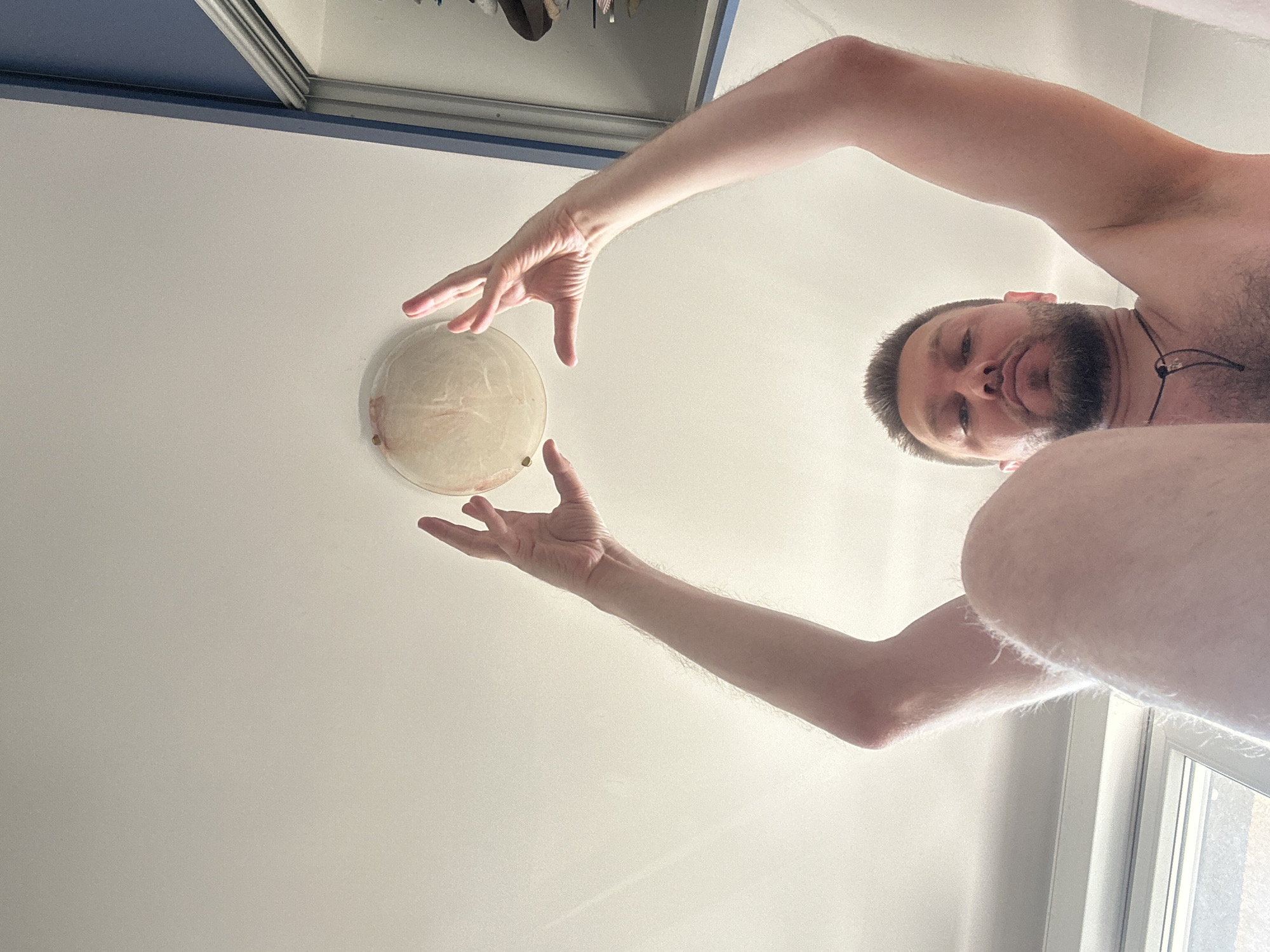 A low-angle, close-up, indoor shot captures a man holding a round, translucent object above his head with both hands. The man has short, dark hair and a beard, and is wearing a dark pendant around his neck. His arms are raised, and his legs are bent, suggesting he is sitting or lying down. The background shows a white ceiling and a glimpse of a blue wall with a closet door. The lighting is bright, indicating it might be daytime. The scene appears to be within a residential setting in Zvezdara Urban Municipality, Serbia.