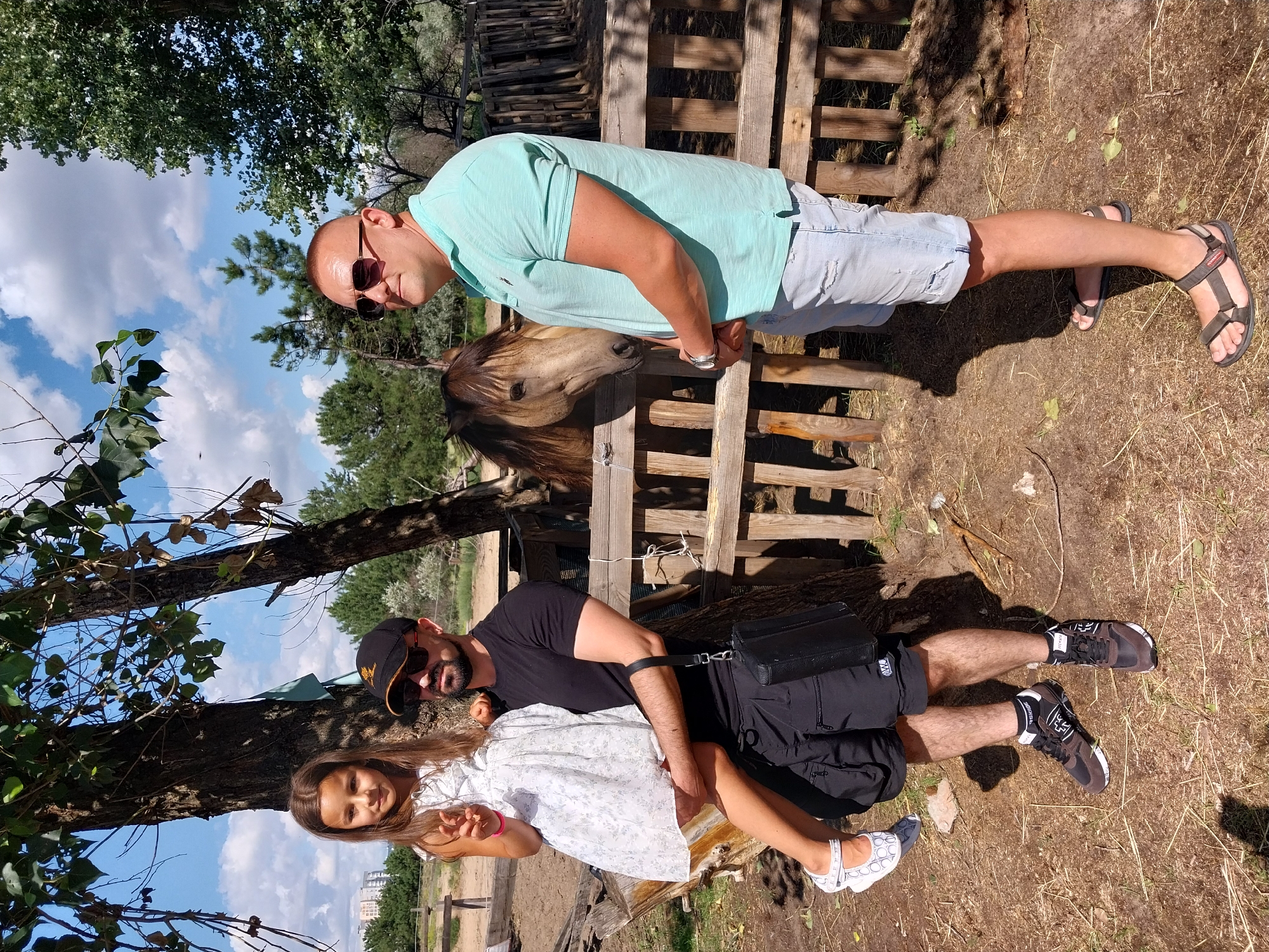 A man carrying a young girl stands in front of a wooden fence at a rural location. The man, on the left, wears a black t-shirt, black shorts, black and brown sneakers, and a black baseball cap. He has a beard and sunglasses. The girl, on the right, is wearing a white floral dress and white sandals. She is being held by the man. To the right of the man, another man, wearing a teal polo shirt and light blue denim shorts, stands with his hands clasped. A horse with a brown mane is visible behind the fence, to the left of the man in the teal shirt. The background includes trees, a blue sky with clouds, and a wooden fence. The ground is covered in dirt and dried grass. The location is in the city of Unknown, Unknown.