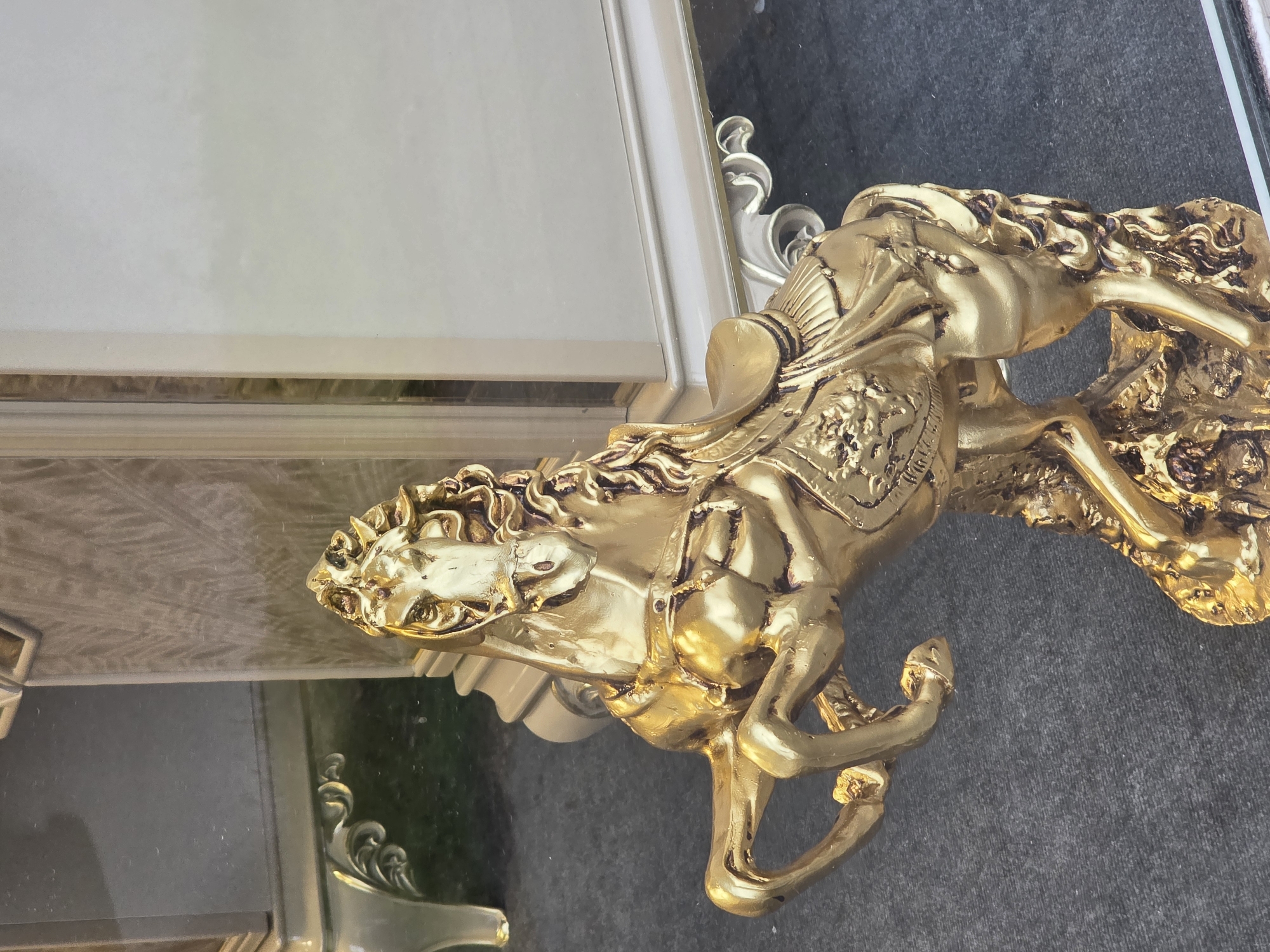 A gold-colored equestrian statue depicting a horse and rider is positioned on a reflective surface, possibly glass, with a gray, textured surface beneath. The statue is highly detailed, with elaborate ornamentation on the horse's trappings and musculature. Part of an ornate white and gold frame is visible near the statue, with its reflective surface indicating an external environment. The specific location in Plovdiv, Bulgaria cannot be determined from the image.