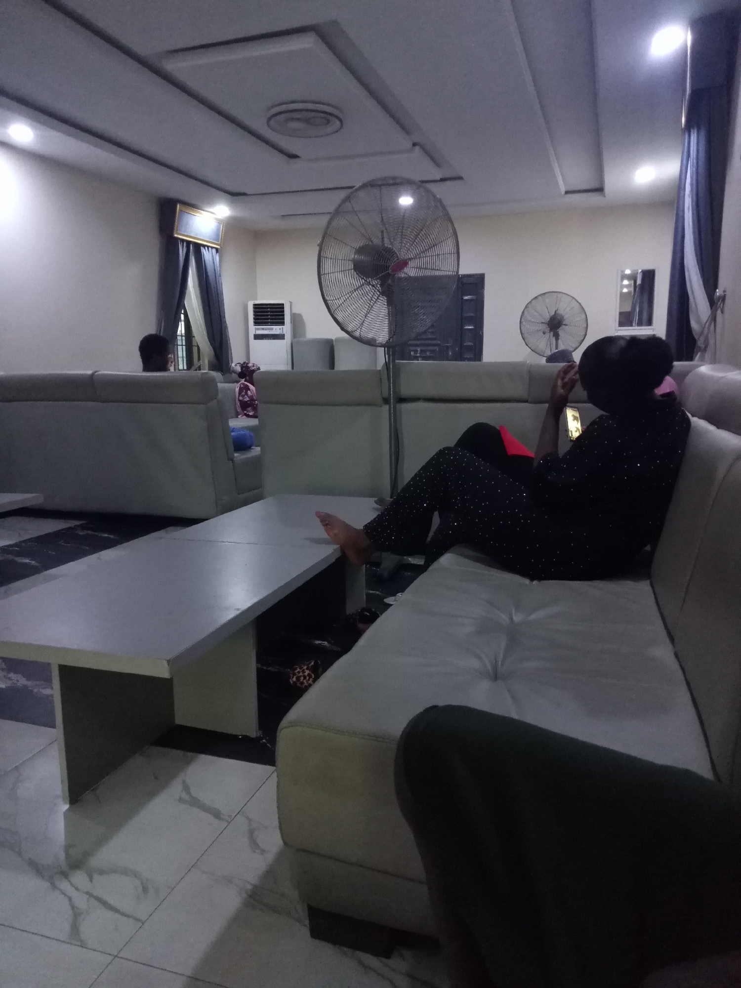 The image depicts an indoor lounge area, likely in Uke, Nigeria, characterized by a suspended ceiling with inset lighting and a fan. In the foreground, a long, light-colored coffee table is visible, with a beige sectional sofa extending to the right. A person, viewed from the back, is seated on the sofa, wearing dark clothing adorned with small white dots and using a mobile phone. Their feet are resting on the coffee table. To the left, another person with dark hair is seated on a separate part of the sofa, facing away from the camera. In the background, an air conditioner unit and another fan are present, along with curtains and a framed picture on the wall. The floor is tiled with a marble-like pattern.