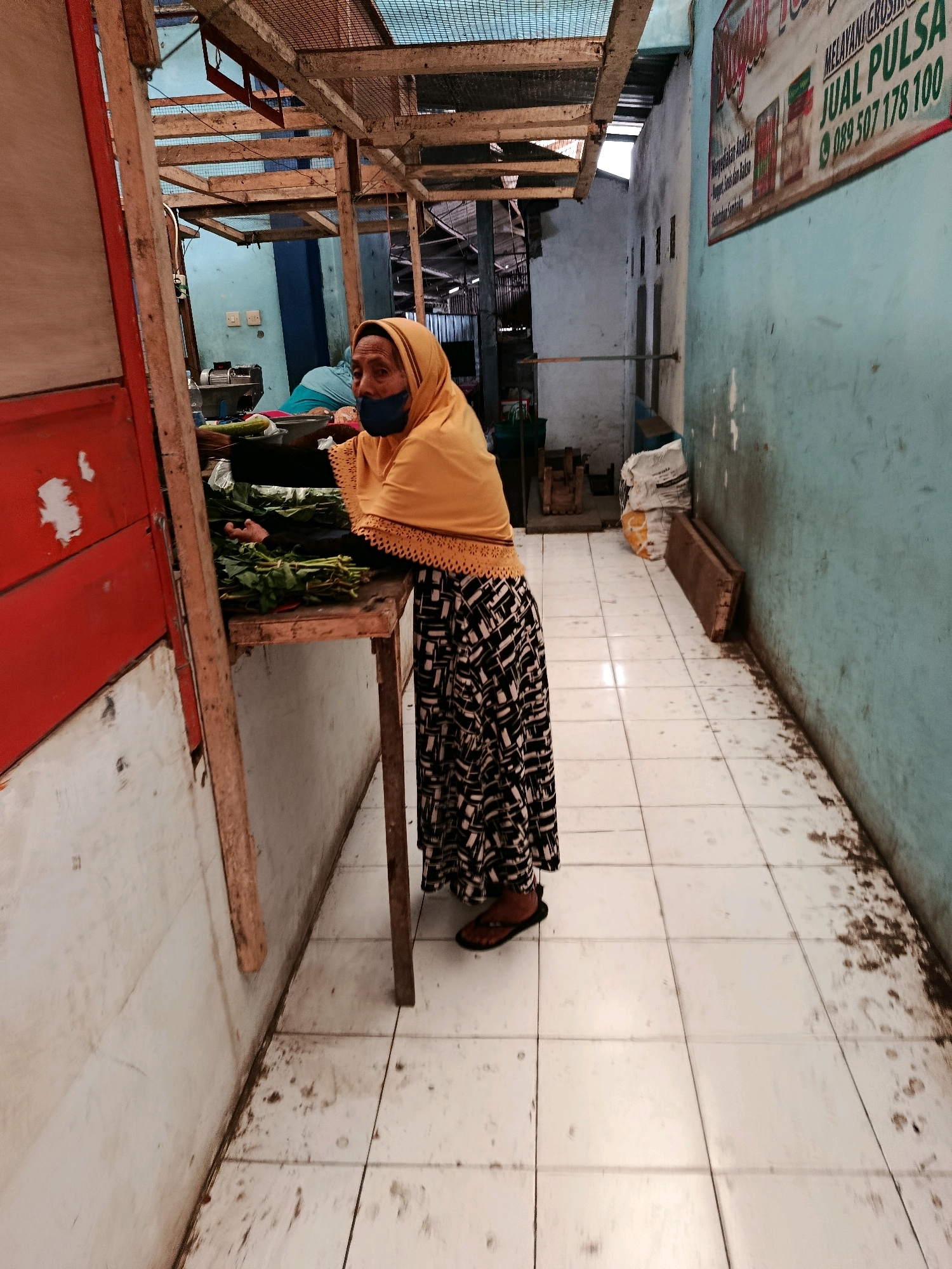 In Bululawang, Indonesia, a woman wearing a yellow headscarf and a face mask stands behind a wooden stall in a market. She is dressed in a black and white patterned skirt and is wearing sandals. In front of her on the stall are bunches of green vegetables. The stall is made of wood and has a red panel on the left side. The floor of the market is tiled with white tiles, which appear dirty. The walls of the market are painted blue and light green. A banner with Indonesian text is visible on the right wall. The overall environment suggests an outdoor or semi-outdoor market setting.