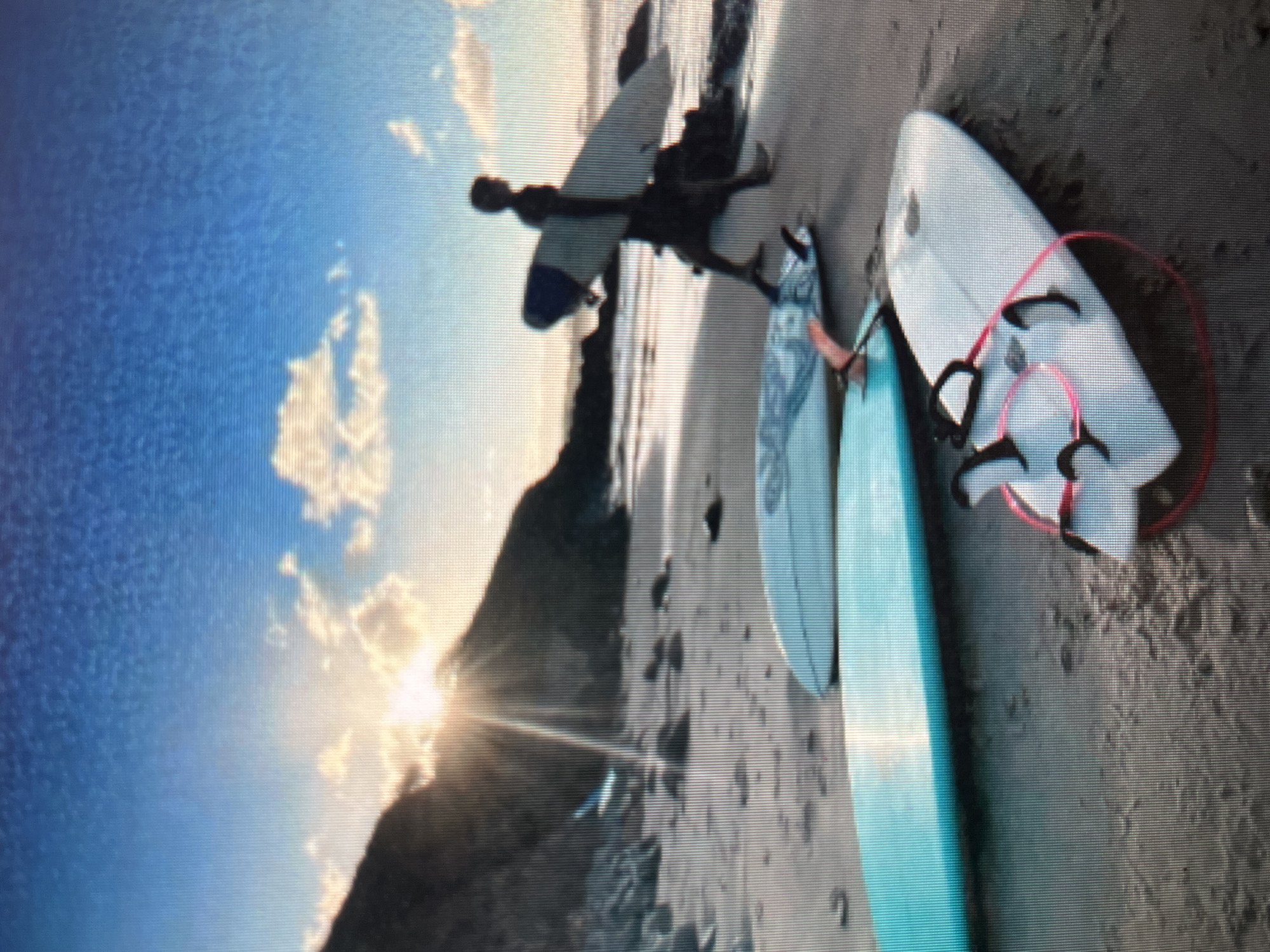 The image depicts a serene beach scene at sunset, or sunrise, in Sejahtera, Indonesia. The low-angle sun casts a warm glow across the sky, illuminating scattered clouds. A silhouette of a person walks along the wet sand, carrying a surfboard. In the foreground, three surfboards are placed on the beach. The closest surfboard is white with black fins and a pink leash. Next to it is a turquoise surfboard. Further back, another surfboard rests on the sand. The background shows a rocky incline leading to the sea. The overall atmosphere is tranquil and evocative of a peaceful coastal environment.