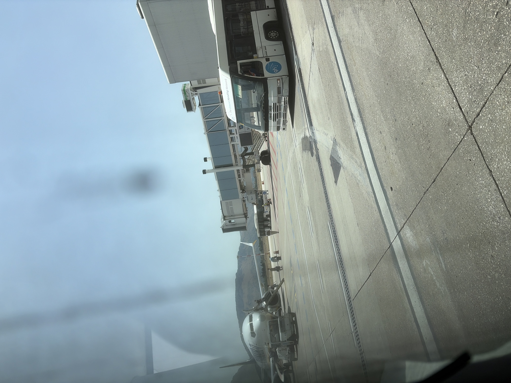 The image captures an airport scene in Cinisi, Italy, likely at Palermo Airport (PMO), with a clear blue sky overhead and distant hills. In the foreground, a paved tarmac is visible, marked with white lines. A white airport bus, identified by the branding "The Ground Handling Network" and "COBUS 3000," is positioned on the right side of the frame, facing left. The bus has tinted windows and a driver is visible inside. To the left of the bus, a jet bridge extends towards a parked airplane. The airplane, a commercial airliner, is white with blue and dark grey accents. A service vehicle, a white tug, is parked near the front of the airplane. Several figures, likely airport personnel, are present on the tarmac, some near the airplane and others near the jet bridge. The overall environment suggests a typical operational day at an airport.