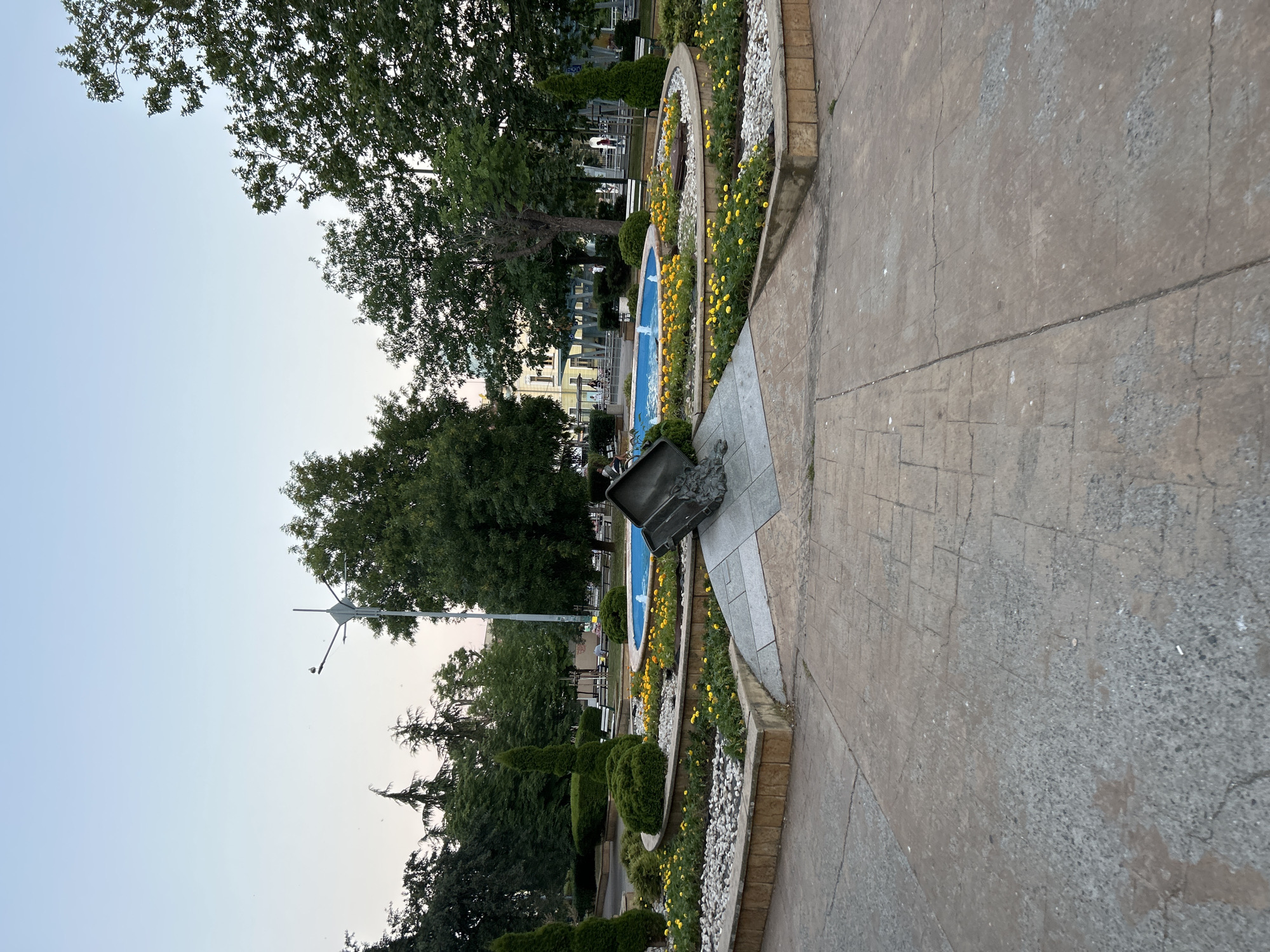 A paved walkway runs diagonally through the frame, leading to a garden bed filled with yellow flowers and white stones. Beyond the garden, a blue fountain with cascading water is visible, surrounded by lush green trees and bushes. In the background, several people are seated at tables, suggesting an outdoor cafe or park setting. A lamp post stands tall on the left side of the image. The sky is clear and light, indicating it is likely late afternoon or early evening. The scene appears peaceful and relaxed, with the natural beauty of the garden and trees creating a serene atmosphere. The overall setting seems to be a public park or a well-maintained outdoor space.