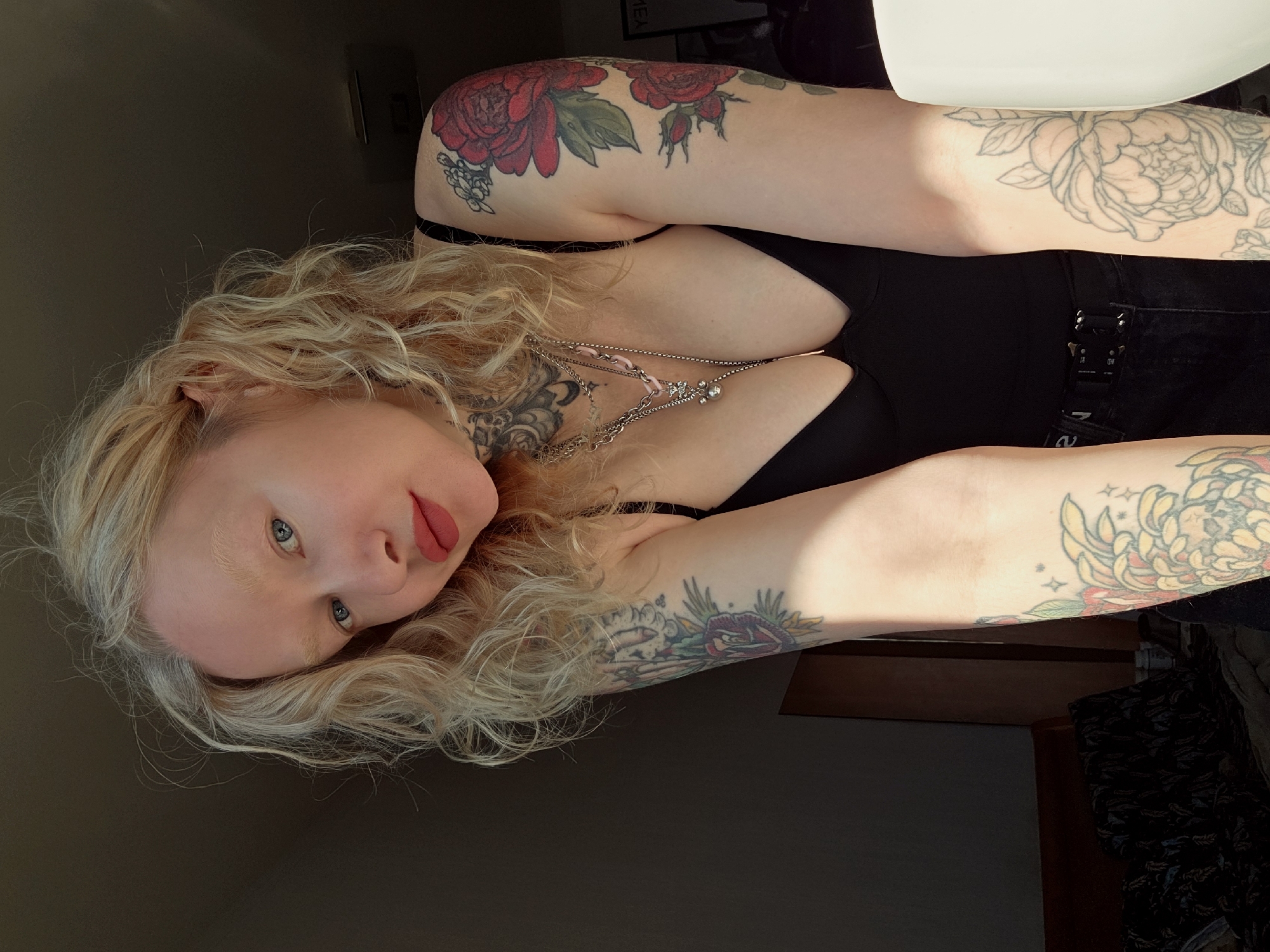 A fair-skinned woman with curly blonde hair and blue eyes is lying down, looking at the camera. She is wearing a black top and jeans with a silver belt. She has multiple tattoos on her arms and neck. The background is dark, and there is a chair visible on the right. There is no indication of the location being Saint Petersburg, Russia.