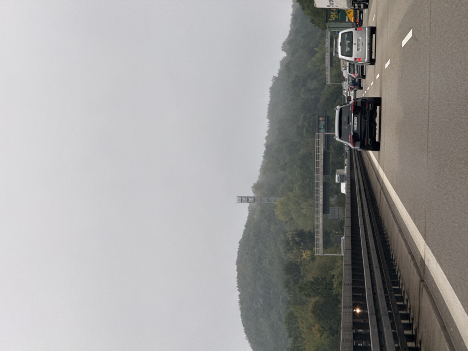 The image displays a multi-lane German autobahn, likely the A8 or A81, situated near Leonberg, Germany, on an overcast day. The perspective is from within a vehicle looking forward, capturing traffic.
In the foreground, the grey asphalt road surface features white solid and dashed lane markings. A metal guardrail with dark vertical posts runs along the left side of the highway.
The midground shows moderate to heavy vehicle traffic. A dark-colored SUV, identified as a Mercedes-Benz with the license plate "BB MB 2774" (Böblingen district), occupies one of the right lanes. To its right, a light-colored van, likely a Volkswagen, bears the license plate "LB C 2307" (Ludwigsburg district). Further ahead, a line of various vehicles, including cars and large trucks, is visible, extending into the distance. An elevated concrete overpass structure with traffic sign gantries spans perpendicular to the highway.
The background is dominated by a densely forested hill. A grey, lattice-style telecommunications tower stands prominently on the upper slope of this hill, partially obscured by atmospheric haze. The sky is uniformly grey and lacks distinct cloud formations. No individuals are explicitly visible, but their presence is inferred by the operation of the vehicles. The scene depicts vehicles in motion or stopped in traffic on a major highway.