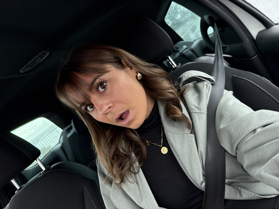 The image depicts a female subject seated in the front passenger seat of a vehicle. The specific location within Villeneuve-Loubet, France, is not discernible from the visual information provided.
**Subject:** A single female with medium-brown hair, styled with bangs and loose waves, is centered in the frame. Her eyes are wide, and her mouth is open in an O-shape, suggesting an expression of surprise or astonishment. She has noticeable eyelashes.
**Attire and Objects:** She is wearing a black turtleneck shirt, over which a light sage-green or pale olive-colored jacket or coat is visible. A gold-toned, chunky link chain necklace with a circular pendant is worn around her neck. Gold-toned, thick hoop earrings are visible on both ears. A black seatbelt is fastened diagonally across her torso and lap.
**Environment:** The subject is situated within a car interior, characterized by black seats with white stitching and a dark ceiling. Through the windows, a blurred exterior environment is visible, displaying light tones and what appears to be water streaks or raindrops on the glass, indicative of precipitation or moisture. The vehicle appears to be stationary or moving slowly.