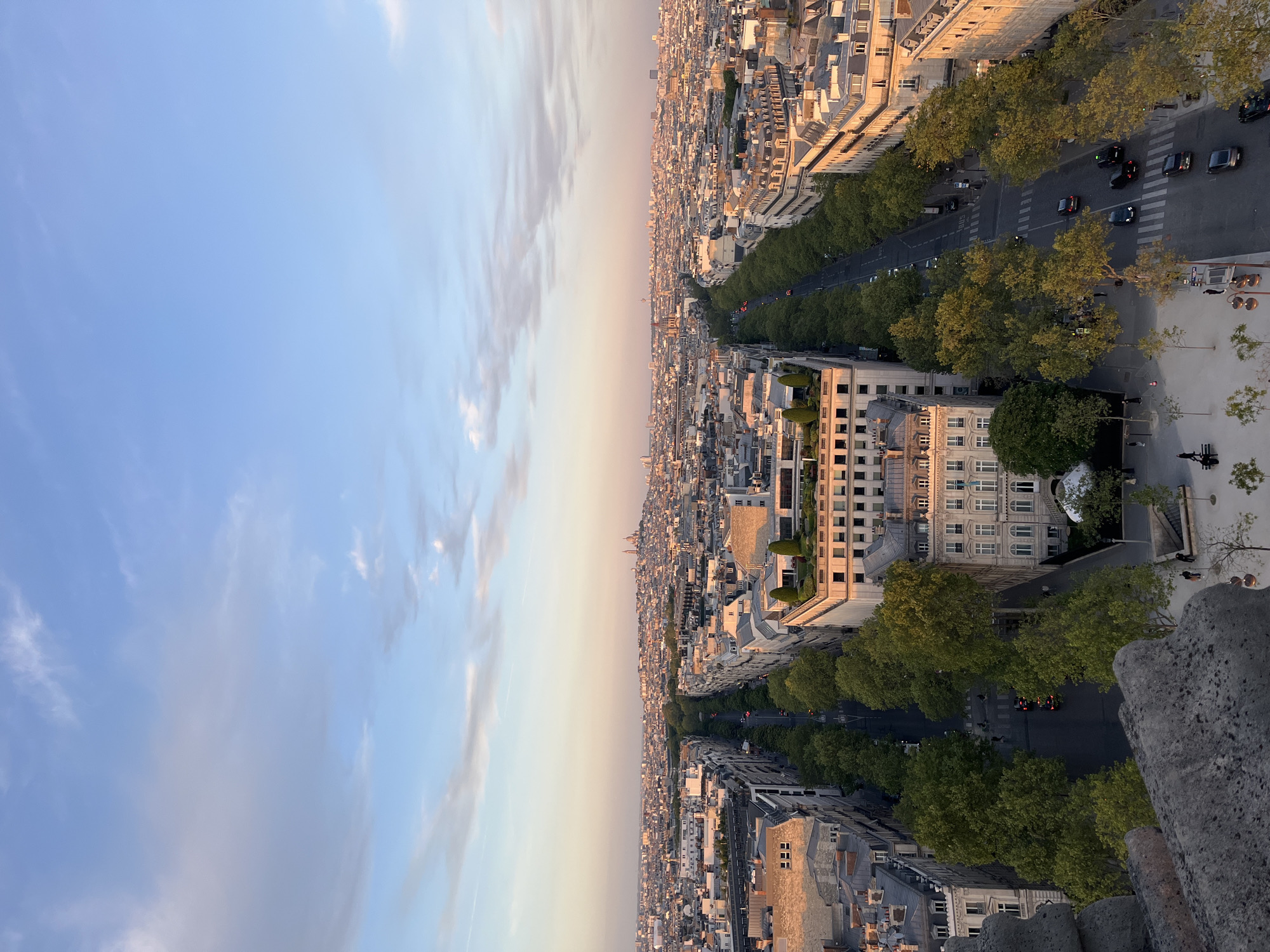 The image captures an expansive, elevated panoramic view of a city, identified as Paris, France, during the late afternoon or early evening, likely at sunset. The scene is dominated by a vast urban landscape characterized by densely packed, uniform Parisian-style buildings. These multi-story structures feature light-colored facades and distinctive grey mansard roofs, many adorned with dormer windows. Some buildings also exhibit balconies or small rooftop terraces with greenery.
A prominent, wide, tree-lined boulevard cuts through the middle ground of the city, extending far into the horizon and dissecting the urban sprawl. Numerous lush green trees form continuous lines along this grand avenue. Several cars are visible on the road, suggesting ongoing city traffic and daily activity. A few tiny figures of people can be discerned on the sidewalks or near the road, though not clearly.
The sky occupies the upper portion of the image, showcasing a gentle gradient from a light blue near the top to a warm, pale orange or pink hue closer to the distant horizon, indicative of a sunset. Scattered wispy clouds add texture to the sky. The buildings are bathed in a warm, golden light, creating long shadows that highlight the time of day. In the far distance, a small dome-shaped structure can be faintly seen, which could be a Parisian landmark. The bottom-right foreground of the image features a rough, textured stone surface, likely part of the vantage point, such as a balcony wall or rooftop edge. No visible text is present. The overall impression conveyed is one of a grand, serene, and historically rich cityscape.