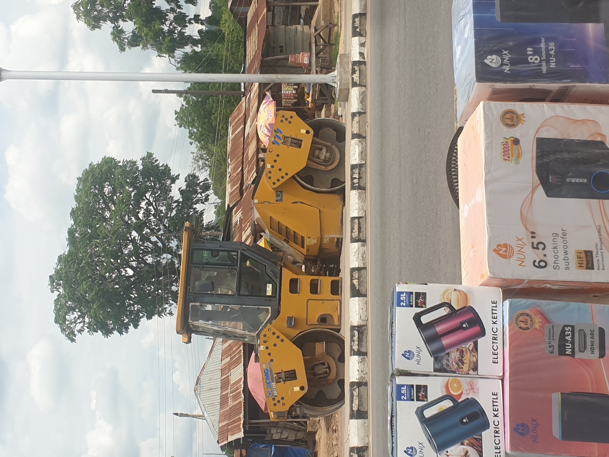 Here is a forensic description of the image.
The image shows a street scene in an unidentified city. A large yellow XCMG XD123S road roller is parked on the side of the road, adjacent to the curb. Next to the road roller is a stack of various products for sale, including Nunix electric kettles and Nunix audio speakers. The items are in their retail packaging. In the background are trees, a street lamp, and buildings with corrugated metal roofs.