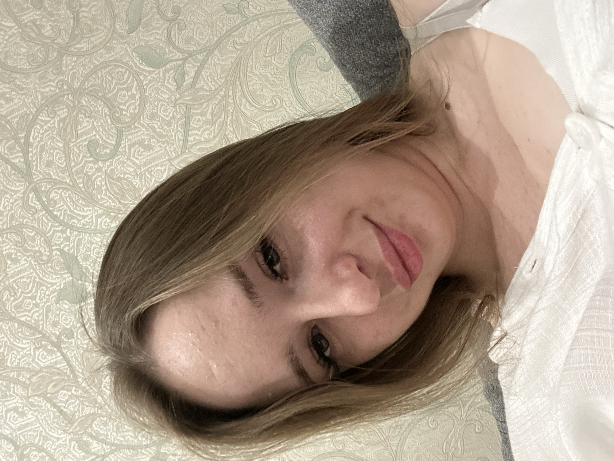 A young woman with light brown hair, fair skin, and light brown eyes takes a selfie in a dimly lit room. She is wearing a white top. The background consists of a patterned wallpaper and a grey fabric. The woman is looking towards the camera with a slight smile. Her facial features are clear, with visible eyelashes, eyebrows, nose, and lips. The lighting in the image is soft, highlighting her face and creating a warm atmosphere. The setting appears to be domestic.