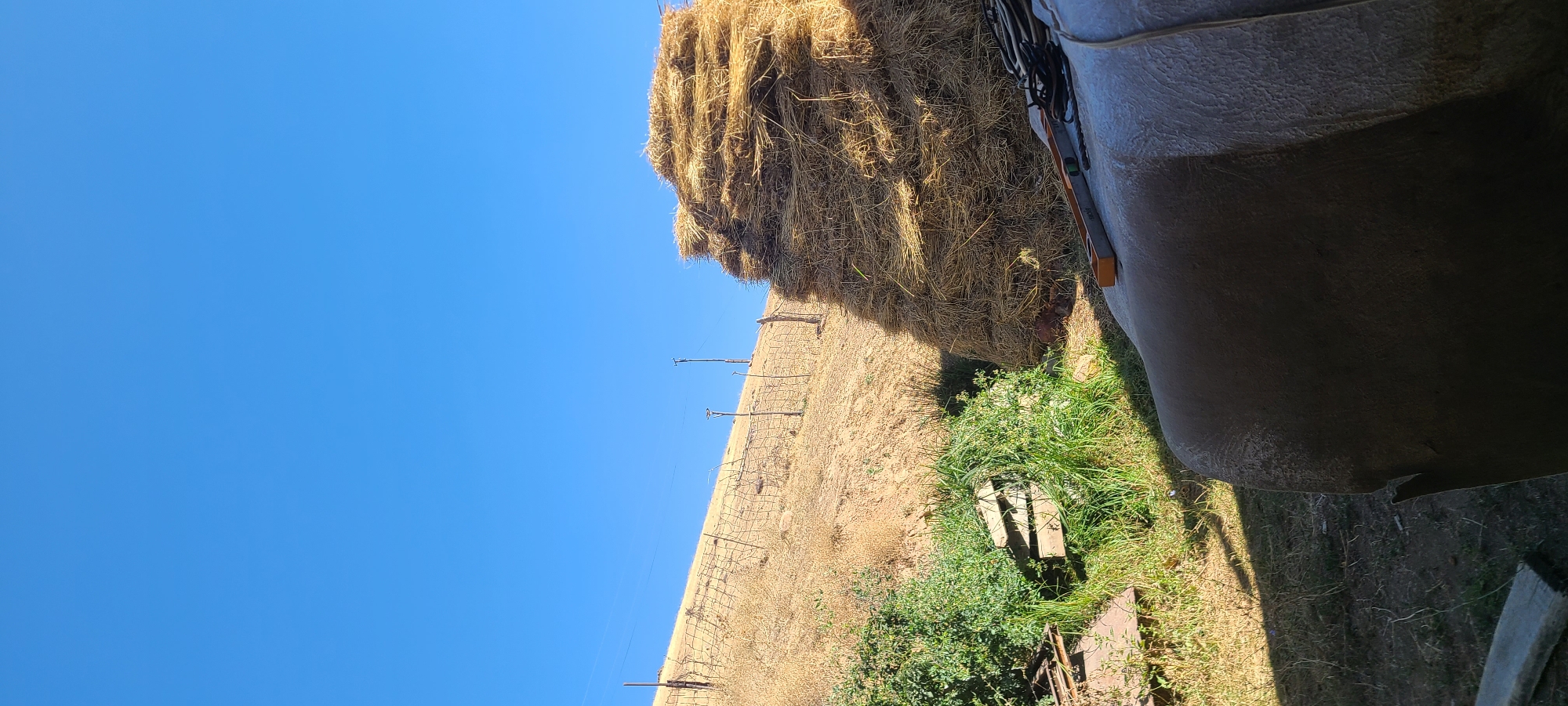 An outdoor scene under a clear blue sky depicts a rural or agricultural setting. In the foreground, a large, light-colored, curved object, possibly a tank or storage container, occupies the bottom right of the frame. On top of this object, a spirit level with orange accents and some coiled dark ropes are visible. To the left of the foreground object, green foliage and some wooden planks are present. Behind this, a dry, grassy slope rises towards the sky, demarcated by a wire fence and a wooden post. Further up the slope, two utility poles with wires stretch across the frame. To the right of the slope, a large, irregular stack of golden hay bales is piled high. The overall impression is one of an outdoor storage area in a dry, possibly arid, environment.